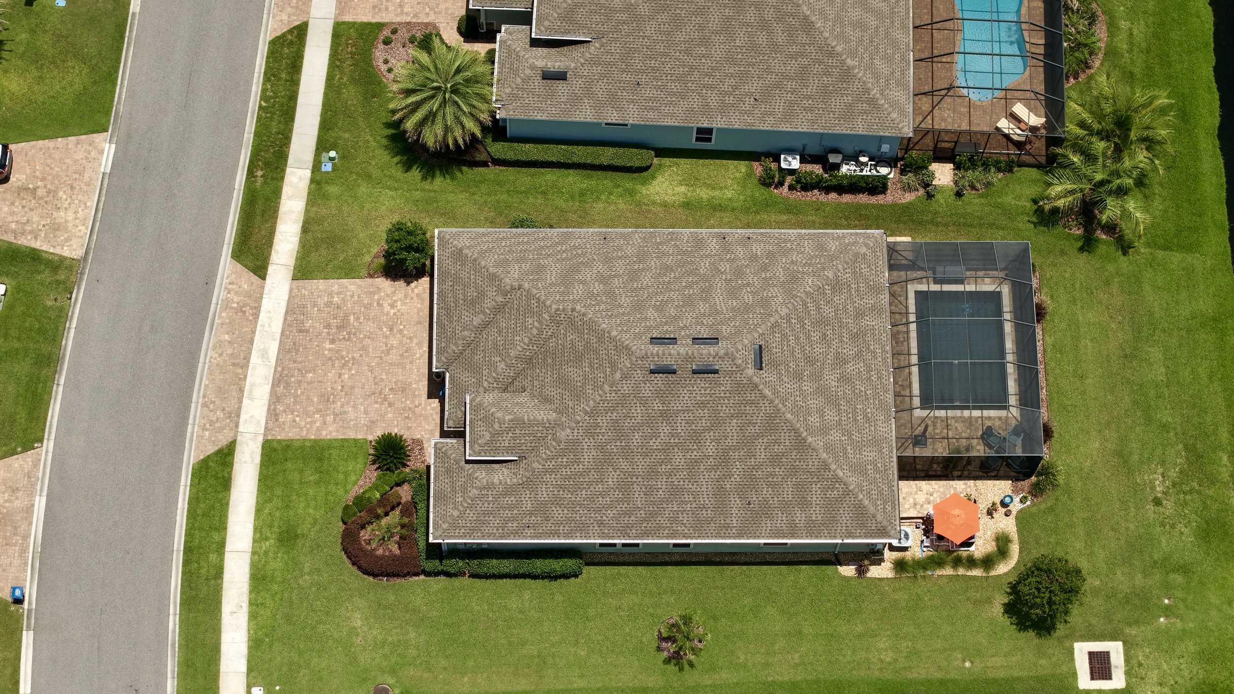 An aerial view of two houses with tiled roofs, surrounded by green lawns, trees, and a paved sidewalk. One house has a screened-in swimming pool with lounge chairs, and a patio with an orange umbrella.