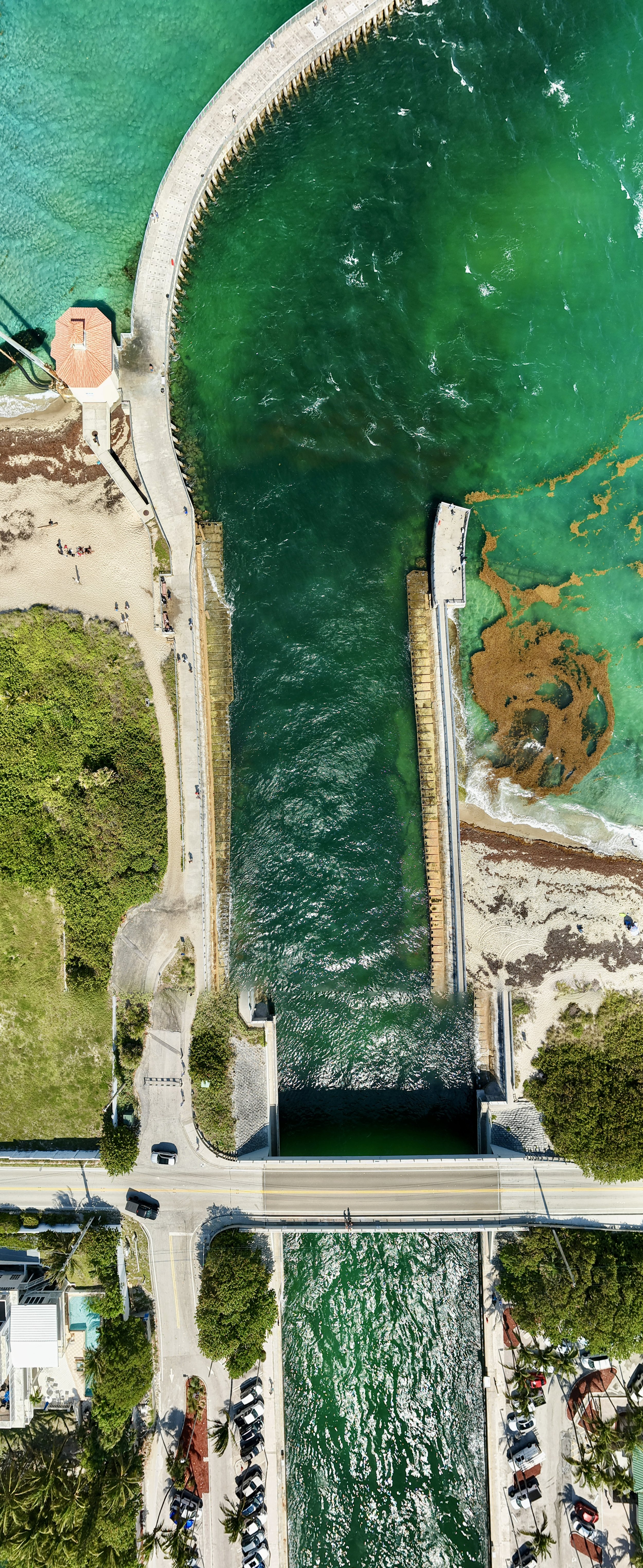 Aerial view of a canal with a dam, surrounded by park area, pathways, trees, and parking lots.