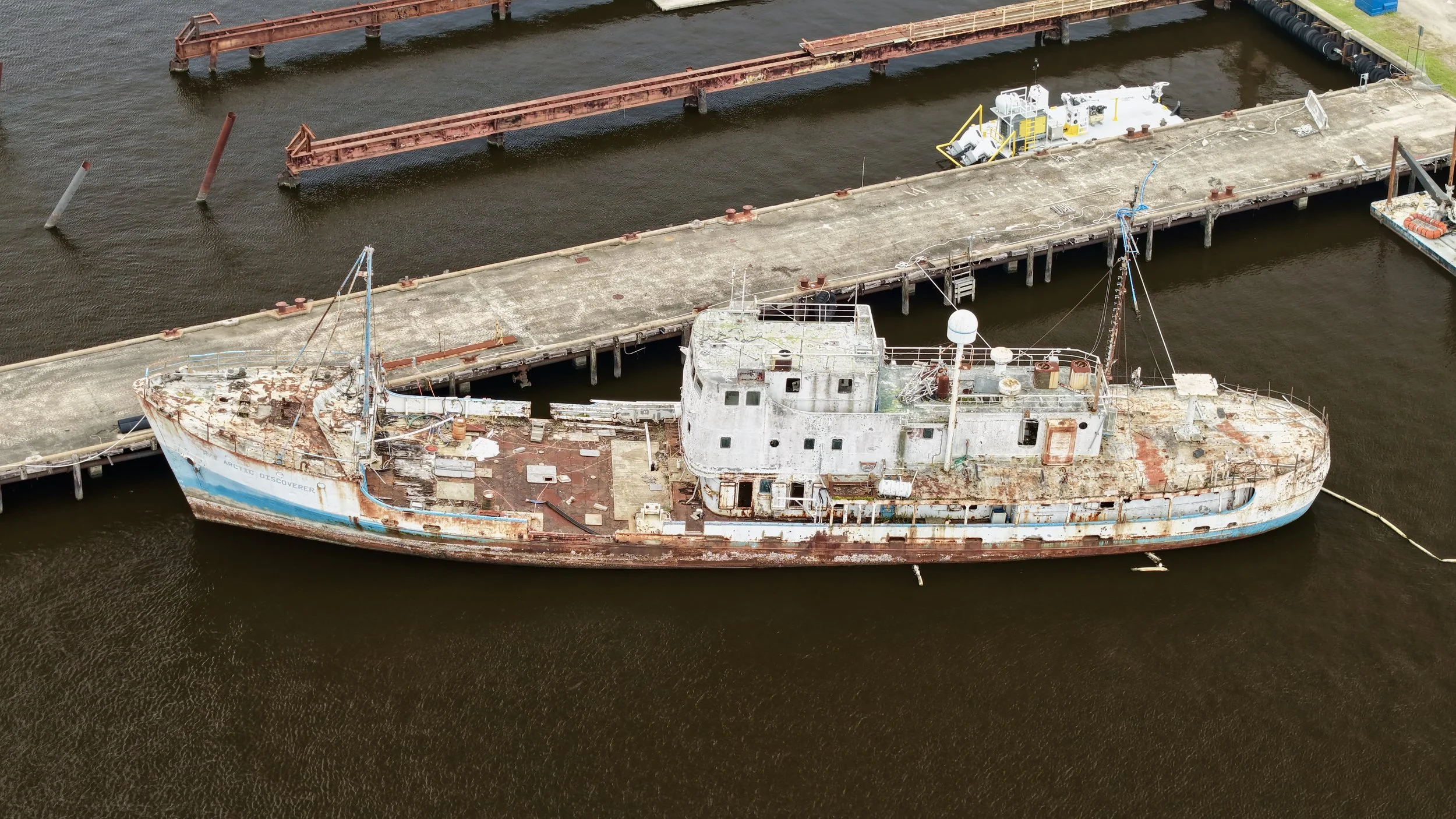 An abandoned, rusty shipwreck docked at a pier with a concrete platform and metal barriers, water around it, and some equipment on the ship.