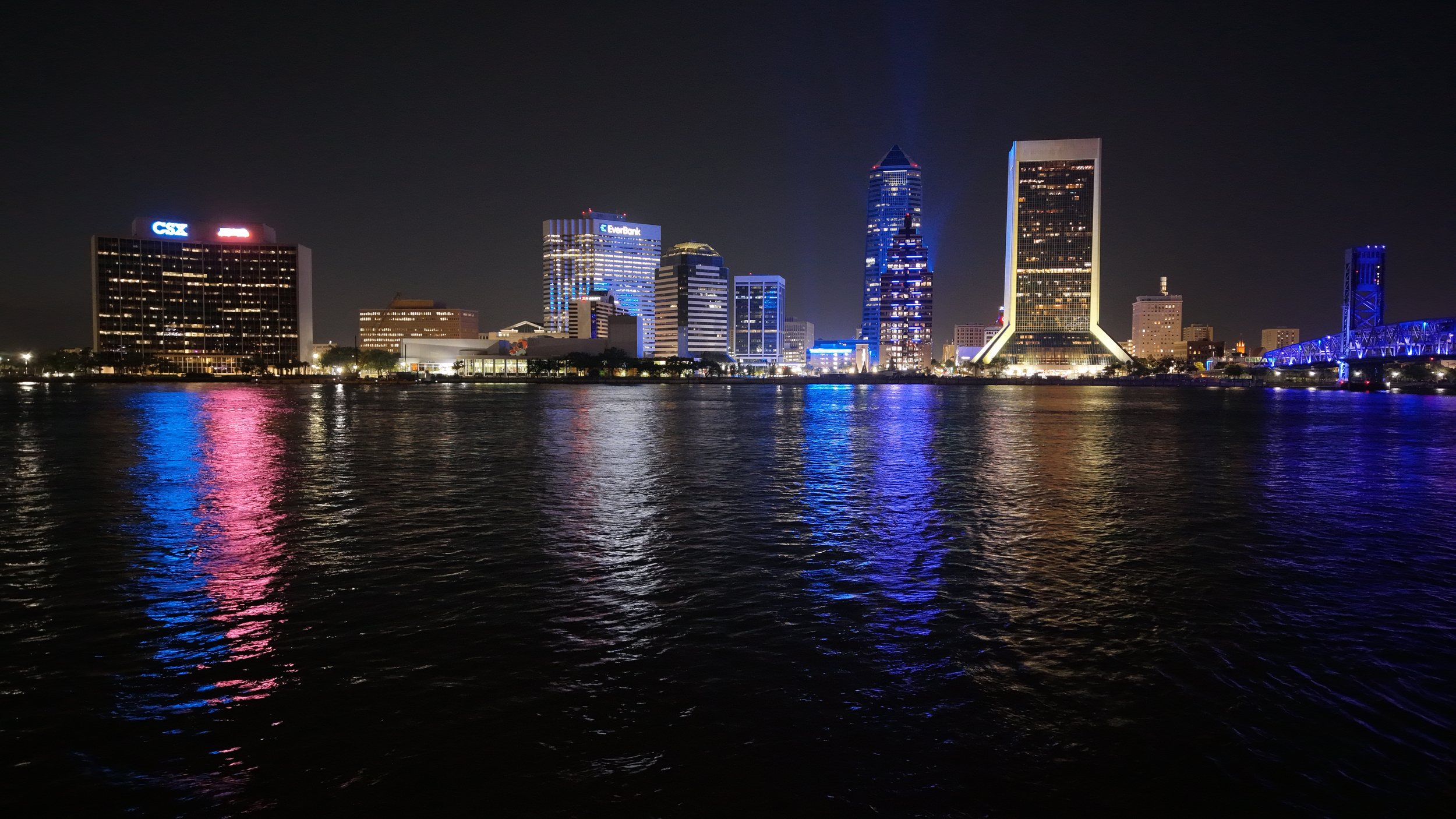 Night view of a city skyline with modern high-rise buildings, illuminated windows, and reflections on water in the foreground.