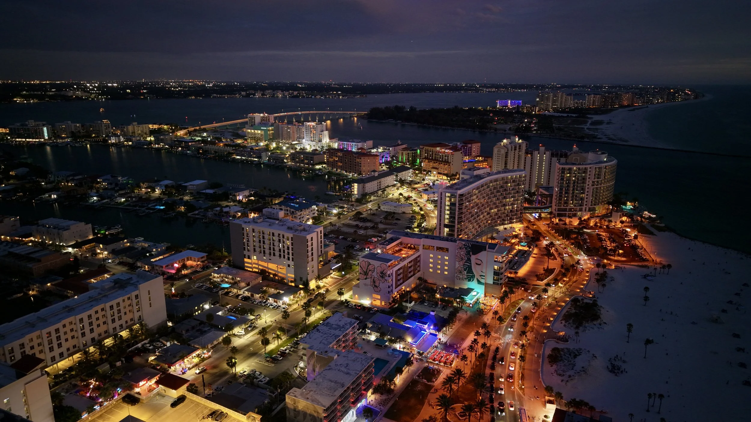 Nighttime aerial view of a city with high-rise buildings, illuminated streets, a waterfront, and a bridge over water, with a dark sky overhead.