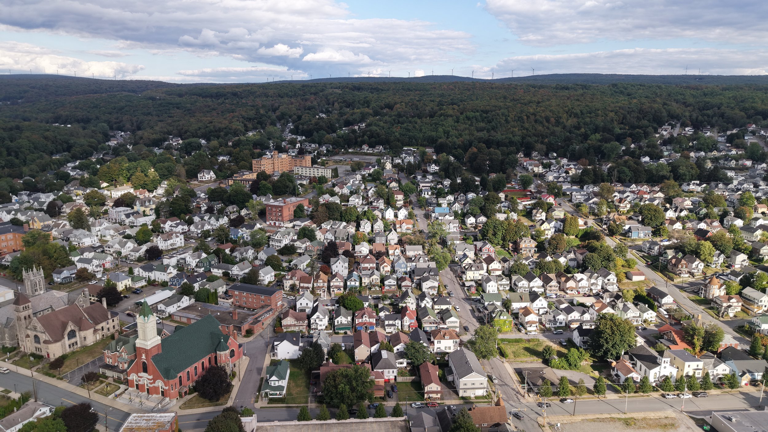 An aerial view of a small town surrounded by green hills and trees with wind turbines on the distant horizon. The town has a mix of residential houses, a church, and a few multi-story buildings.