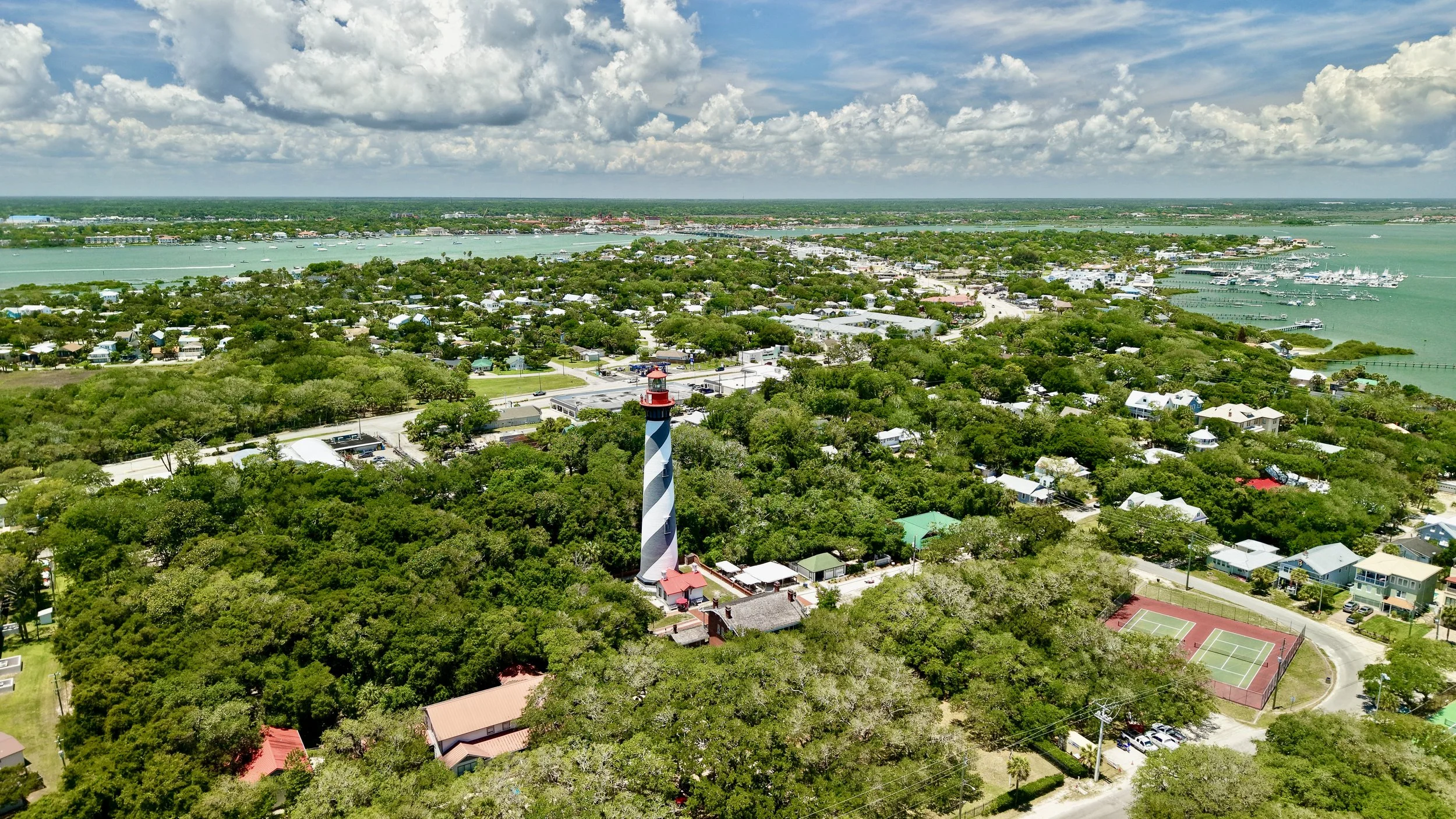 Aerial view of a coastal town with a mix of residential homes, a lighthouse, a marina with boats, and green trees under a partly cloudy sky.