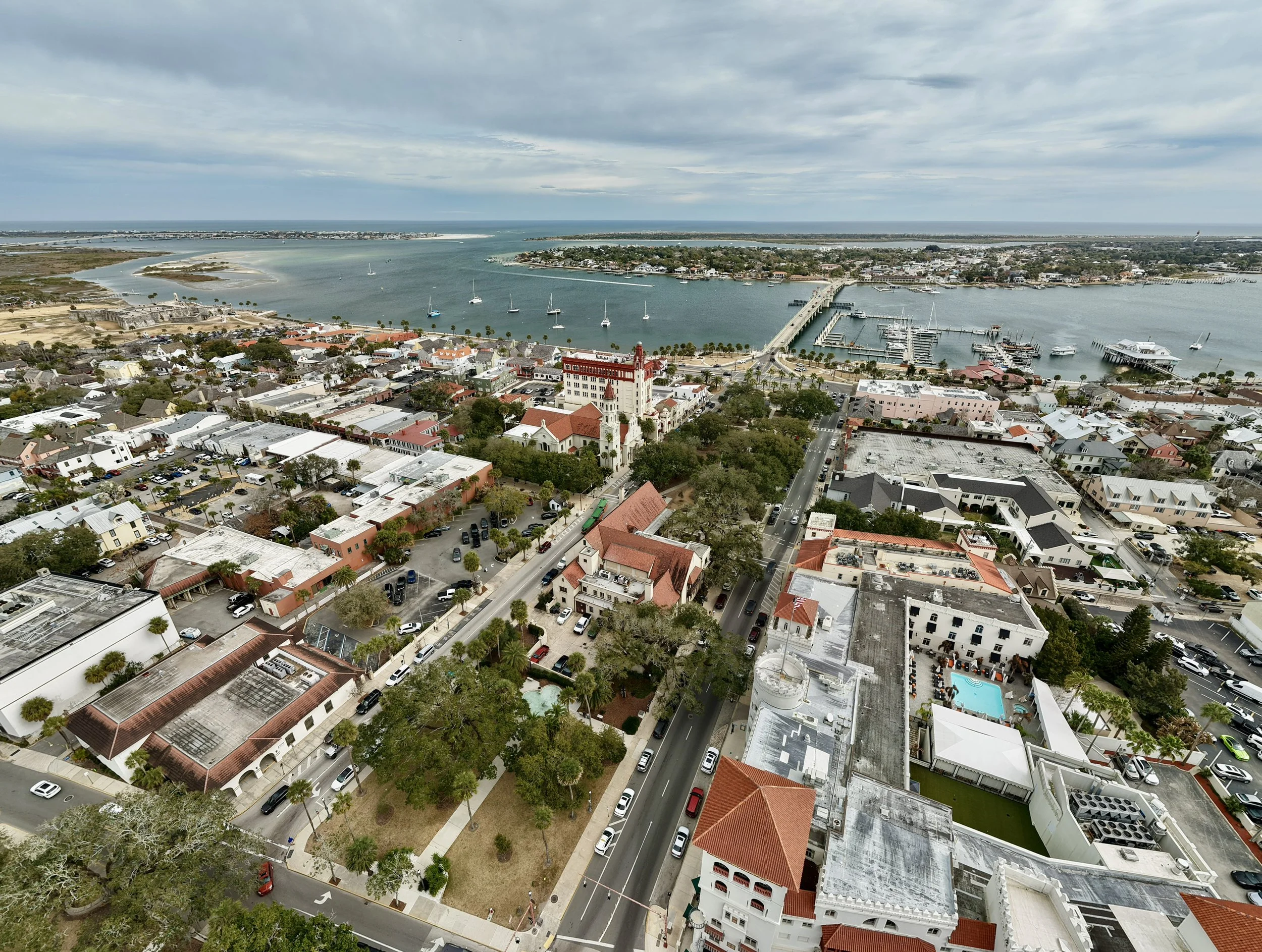 An aerial view of a coastal town with a harbor, sailboats, and a mix of modern and historic buildings, with water and islands in the background.