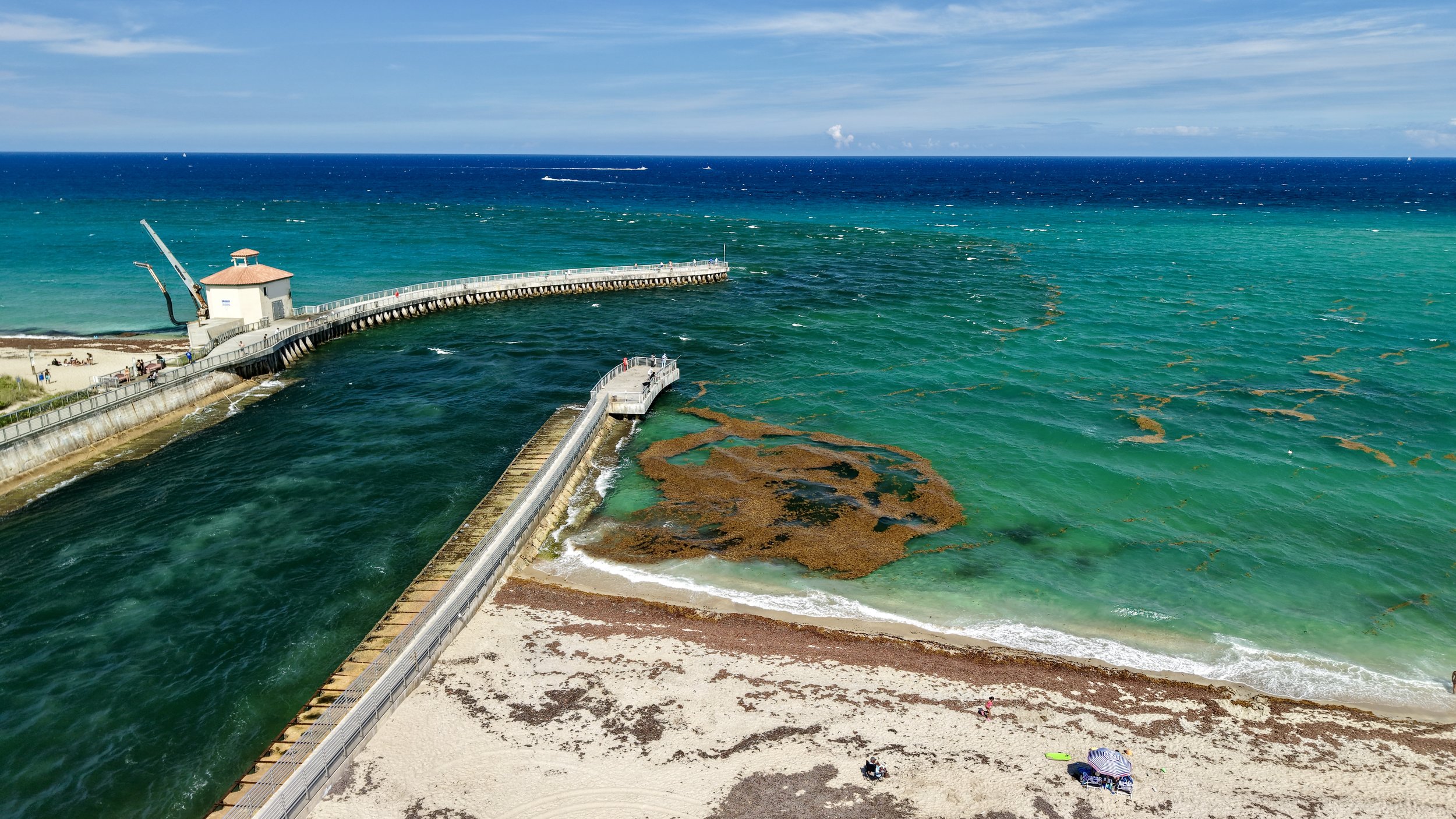 Aerial view of a pier extending into the ocean near a sandy beach with people under umbrellas, with turquoise water and a cloudy sky.