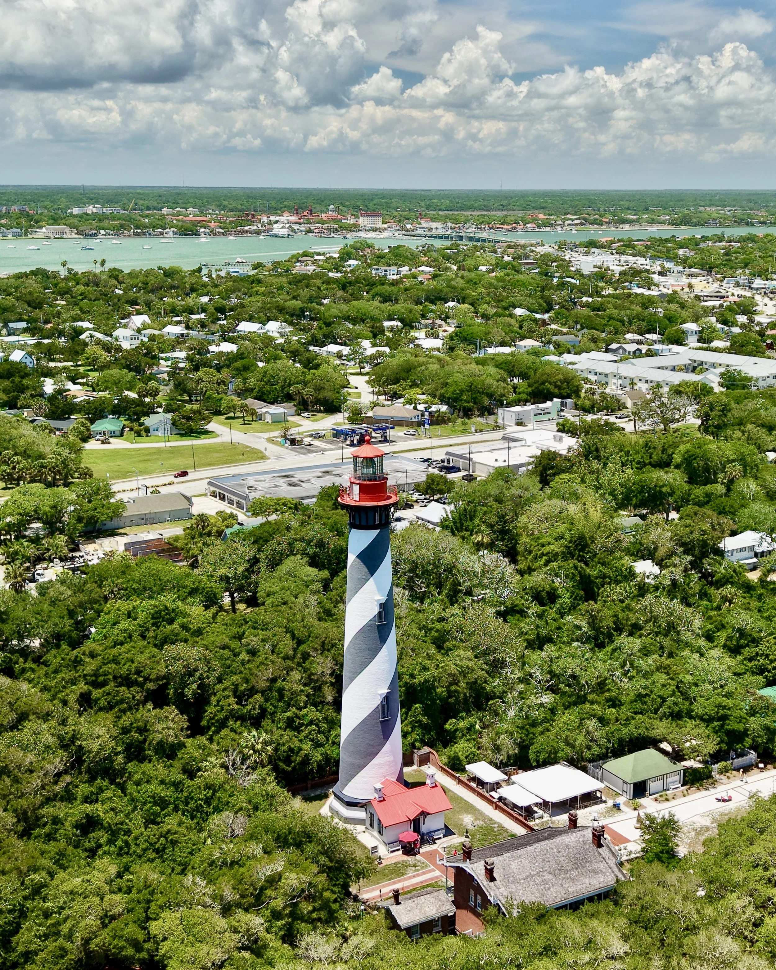A coastal town with a lighthouse in the foreground, residential homes surrounded by trees, a body of water with boats, and a city skyline with buildings across the water under partly cloudy skies.