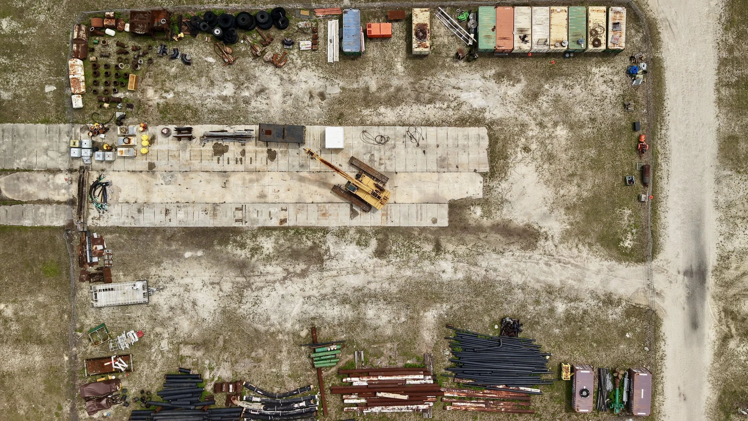 An aerial view of an outdoor yard with scattered industrial equipment, tires, traffic cones, metal pipes, and containers, with a gravel and concrete ground.