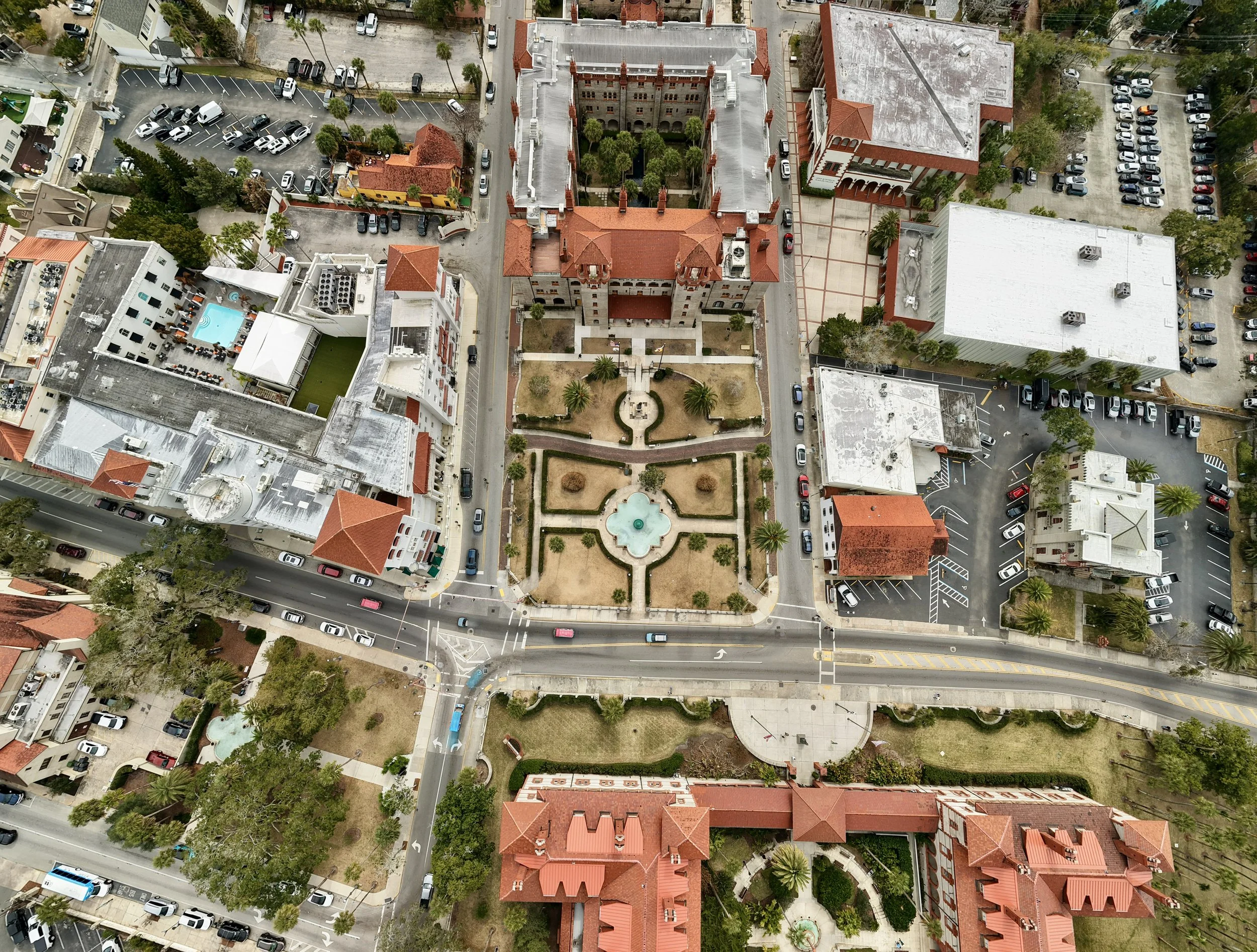 An aerial view of a city block featuring a large historic building with red tile roofs, surrounded by parking lots, smaller buildings, trees, and streets with cars.