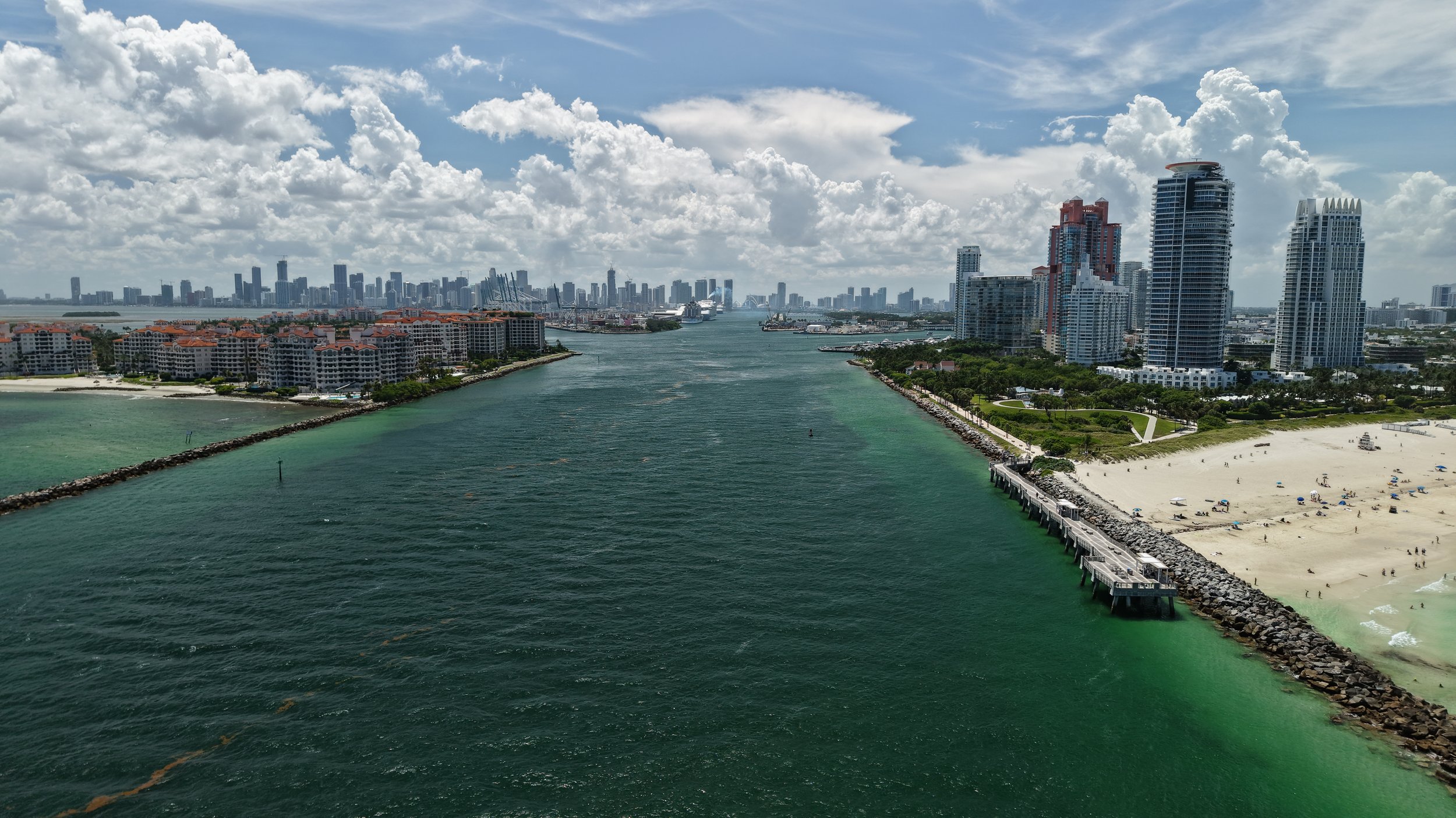 Aerial view of a coastal city with tall buildings, a sandy beach with umbrellas, and a body of water separating different parts of the city under a partly cloudy sky.