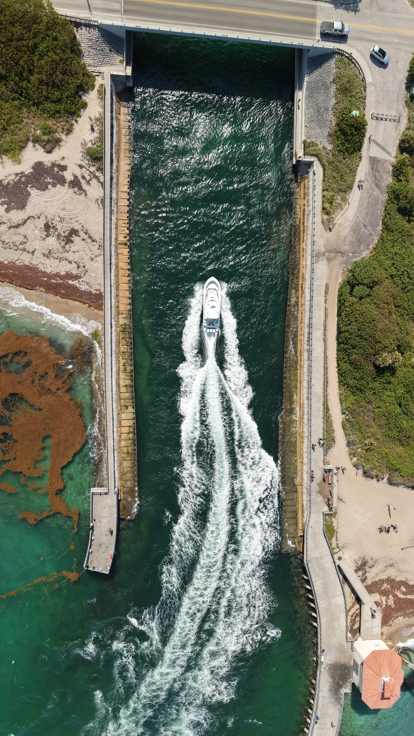 An aerial view of a canal with a white boat moving through, creating wake waves. The canal is flanked by walkways, greenery, and a small sandy beach area with some brown seaweed on the left.