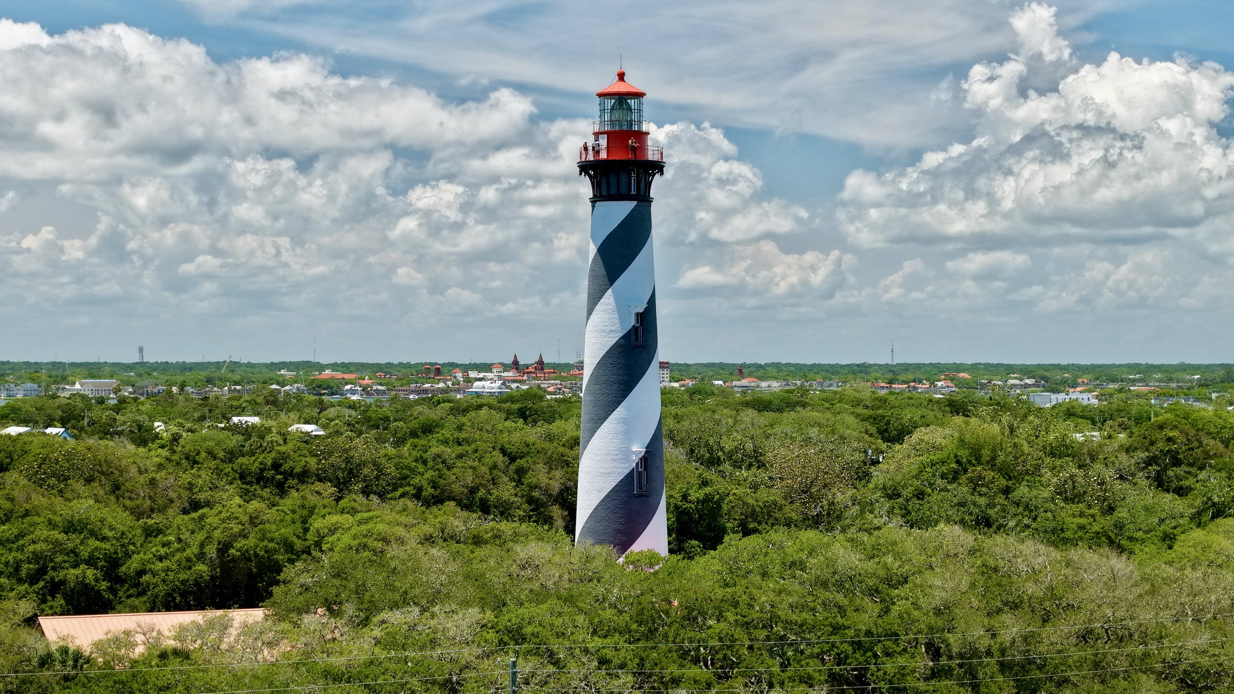 A lighthouse painted with black and white spiral stripes stands over green trees with a town and cloudy sky in the background.