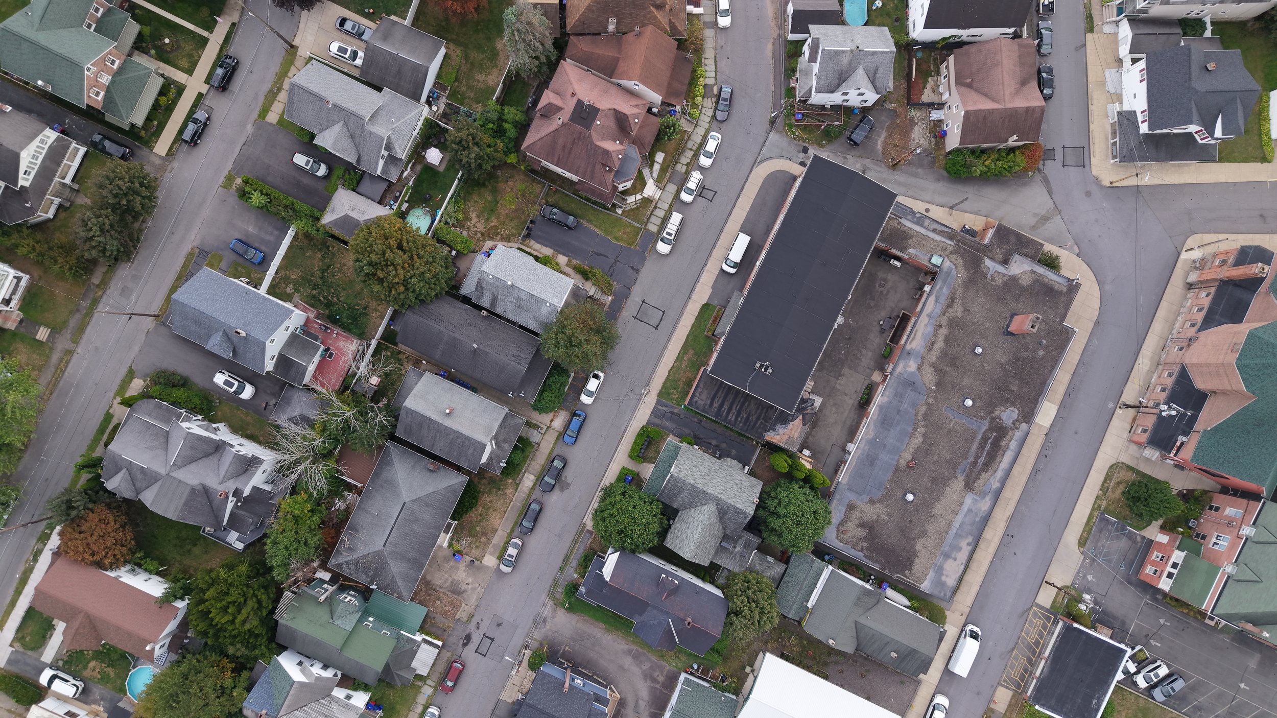 An aerial view of a residential neighborhood with multiple houses, parked cars along the streets, trees, and a few backyards, some with ponds or pools, and streets intersecting at corners.