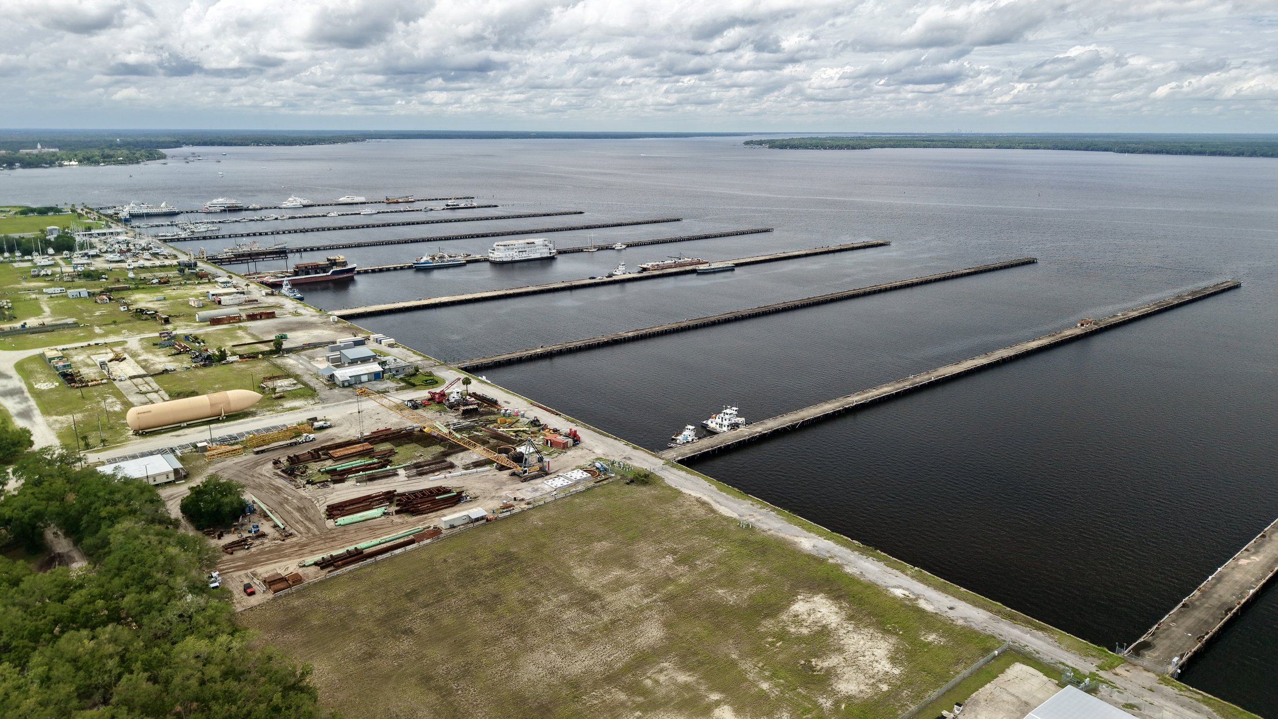 Aerial view of a marina with multiple docks, boats, and ships on a large body of water, adjacent to an industrial area with equipment, storage tanks, and construction materials.