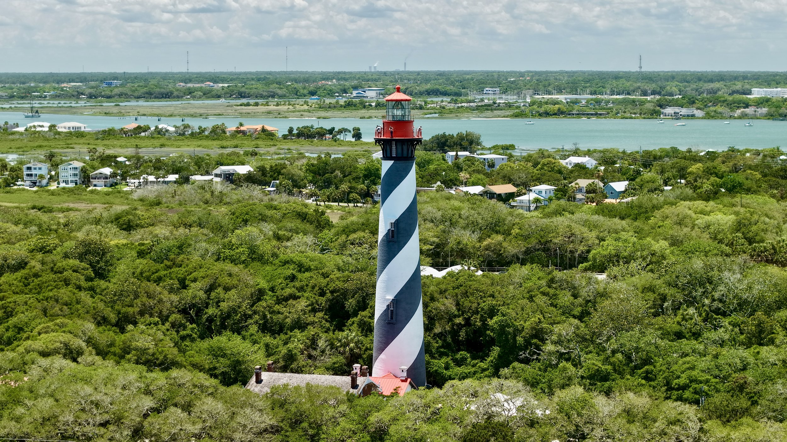 A black and white striped lighthouse with a red top standing among green trees, overlooking a river with several boats, with buildings and a distant shoreline in the background.