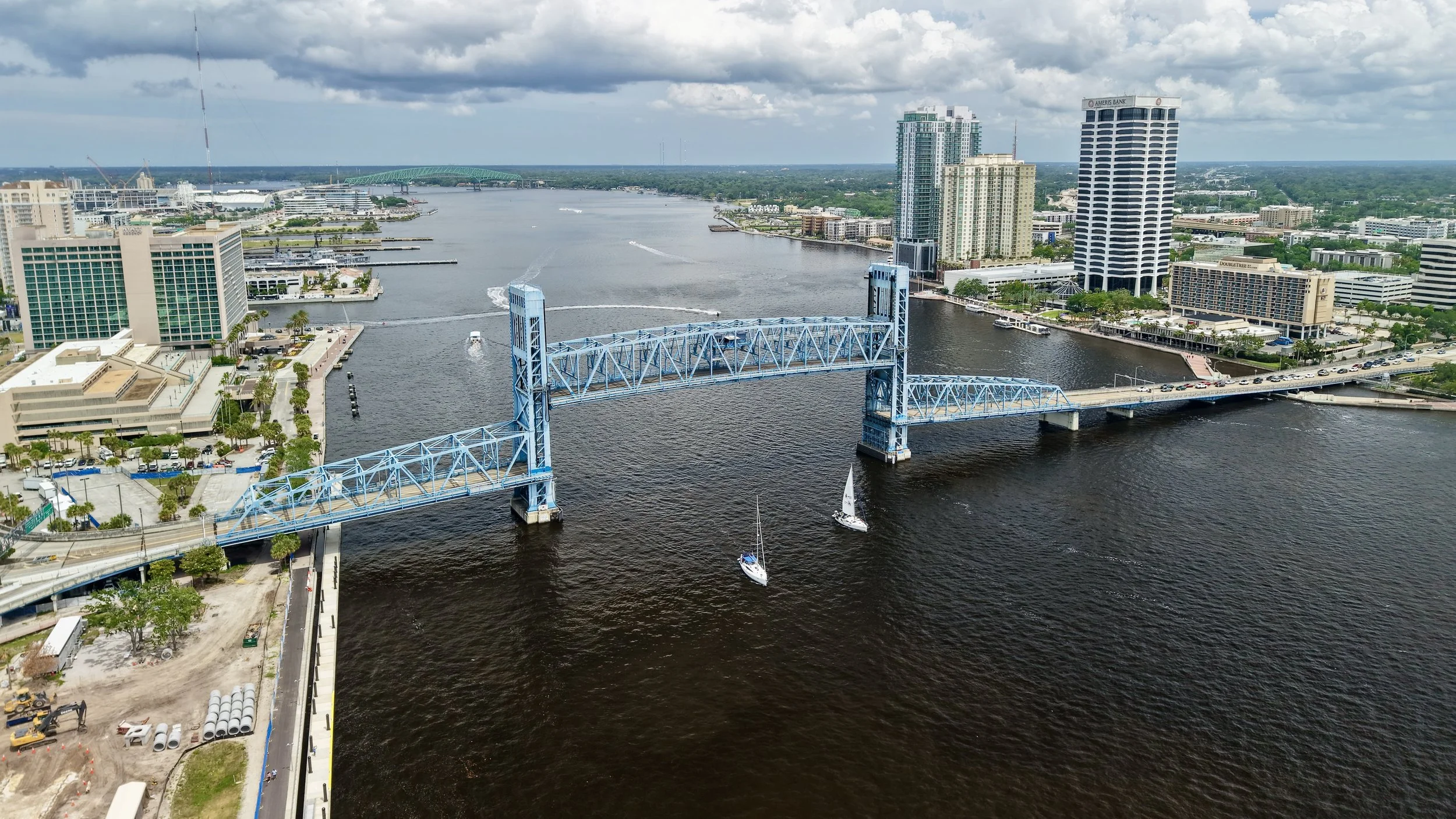 Aerial view of a city with a blue bridge crossing a river, surrounded by high-rise buildings, boats on the water, and a partly cloudy sky.