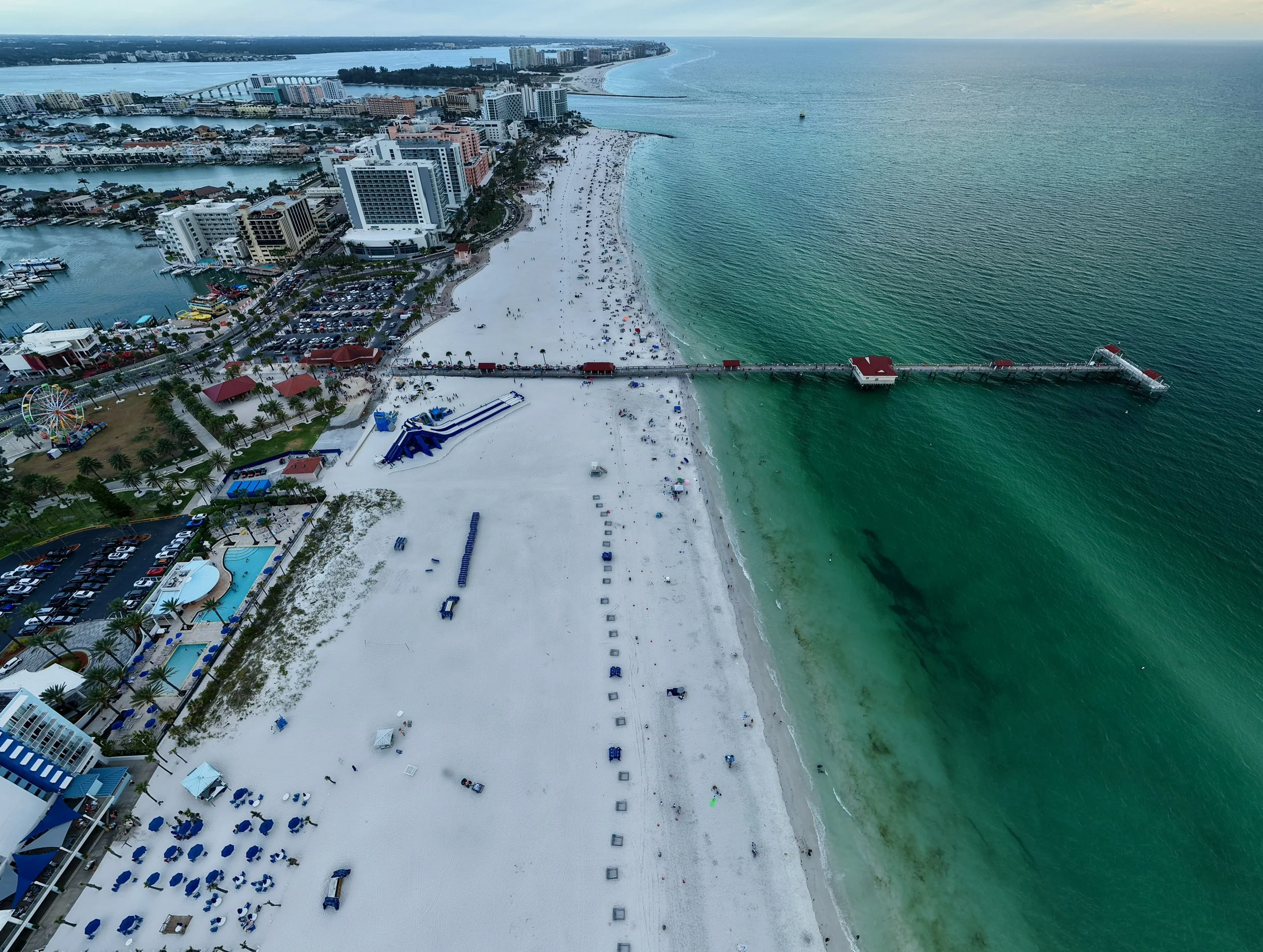 Aerial view of a beach with white sand and turquoise water, lined with umbrellas and lounge chairs, and a pier extending into the ocean. There are high-rise buildings and a marina nearby, with a ferris wheel and playground in the vicinity.