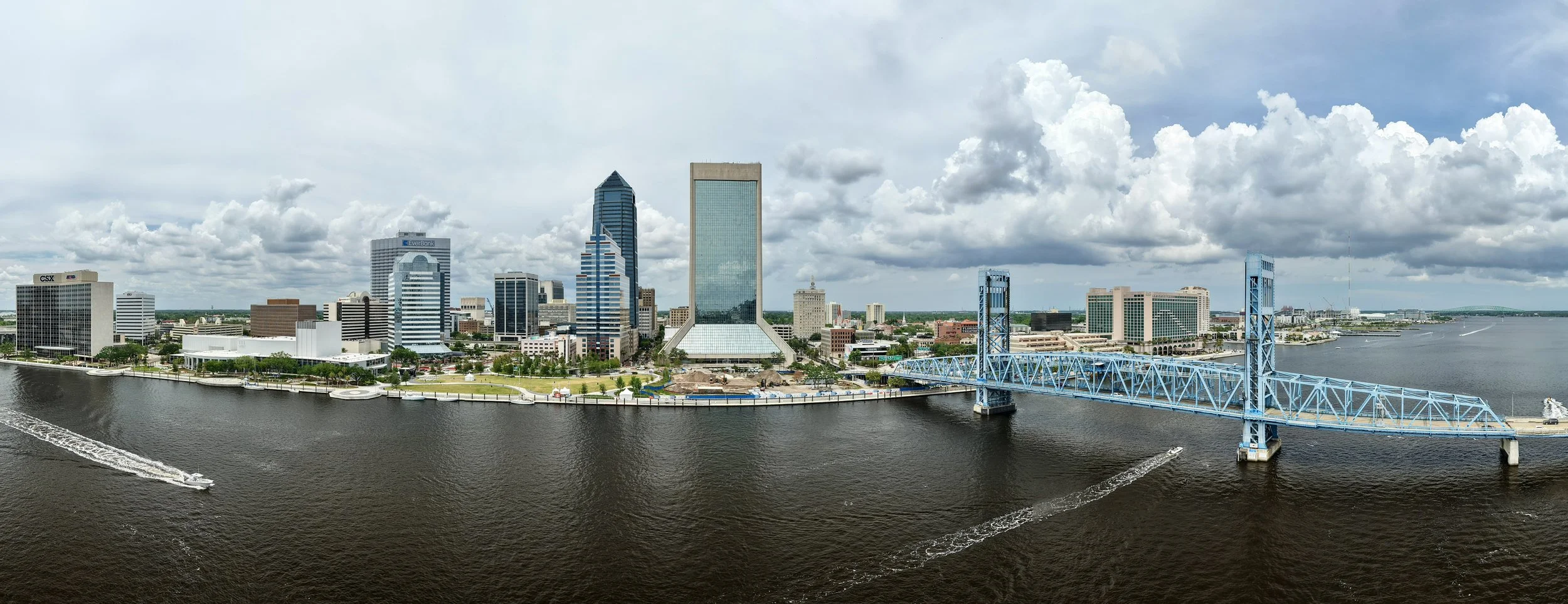 A city skyline with tall modern buildings along a river, featuring a prominent blue bridge and several boats on the water. Clouds are scattered across the sky.