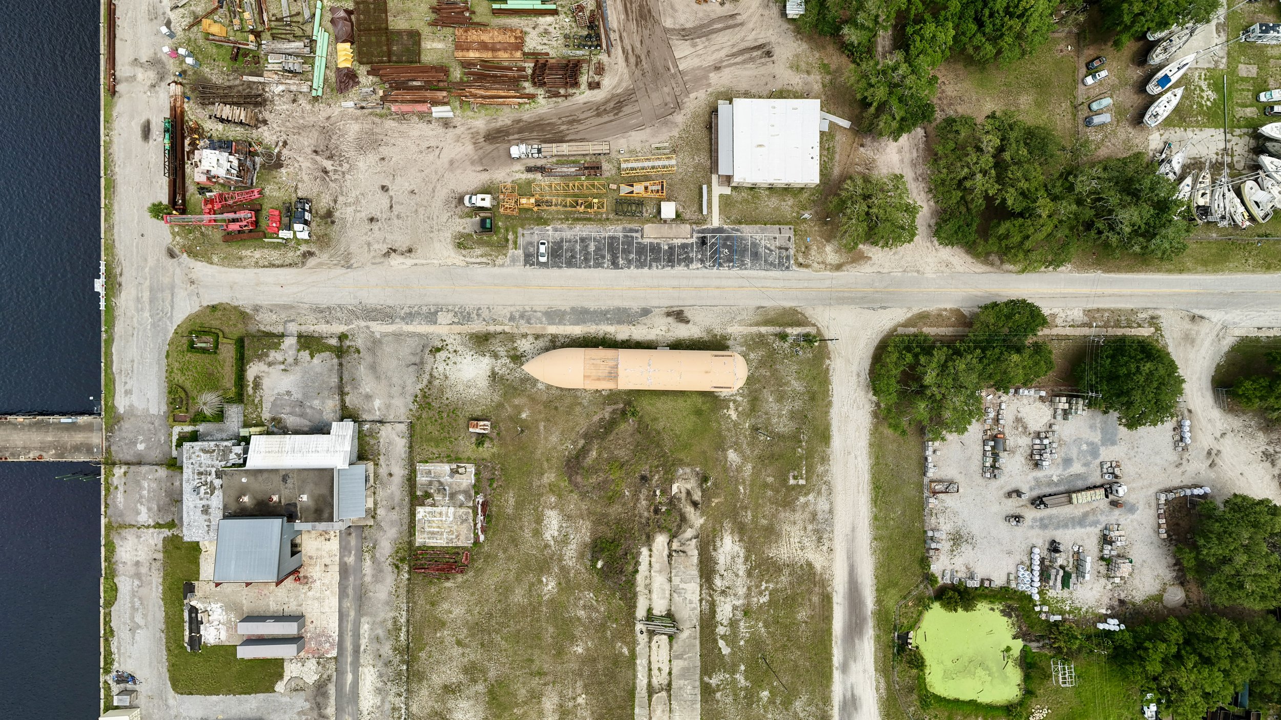 An aerial view of an industrial area with boat salvage yard, storage areas for equipment and materials, parking lot, trees, and nearby water.