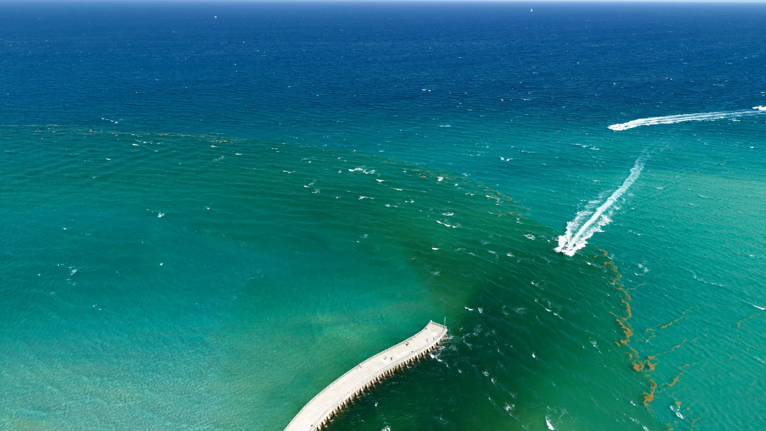 Aerial view of a curved pier extending into turquoise ocean water with several boats and wake trails visible.