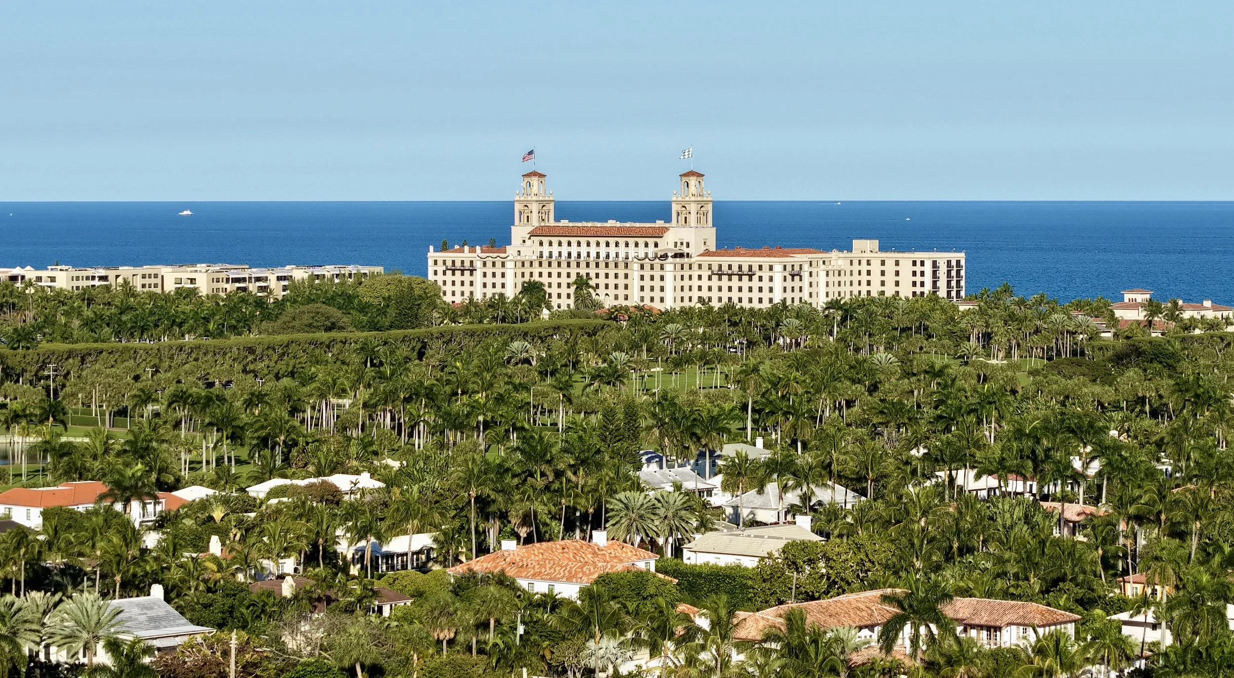 View of a coastal hotel with two towers and flags, surrounded by lush green trees, overlooking the ocean on a clear day.