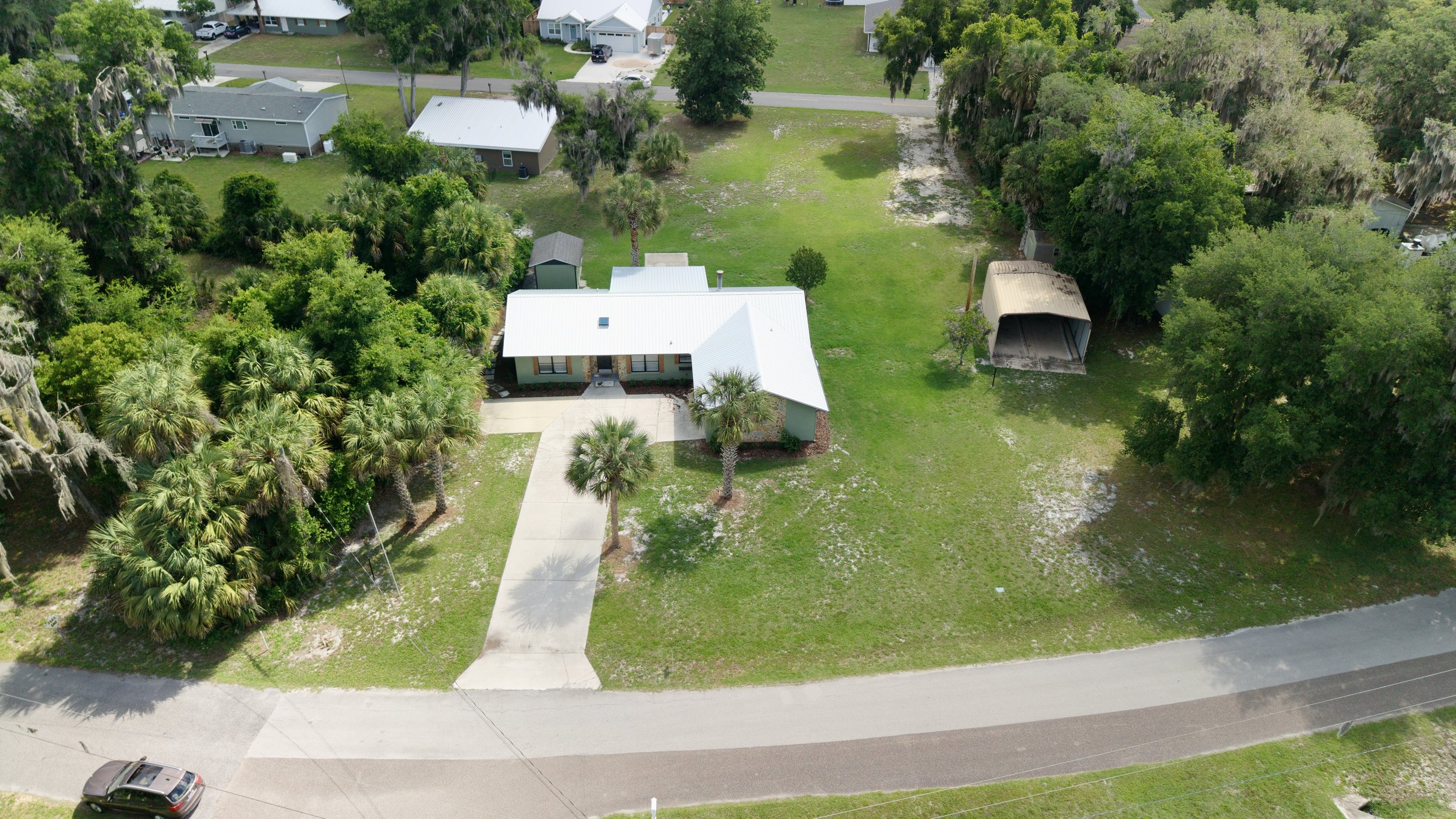 Aerial view of a suburban house with a white metal roof, driveway, backyard, and surrounding greenery, including palm trees and other trees.