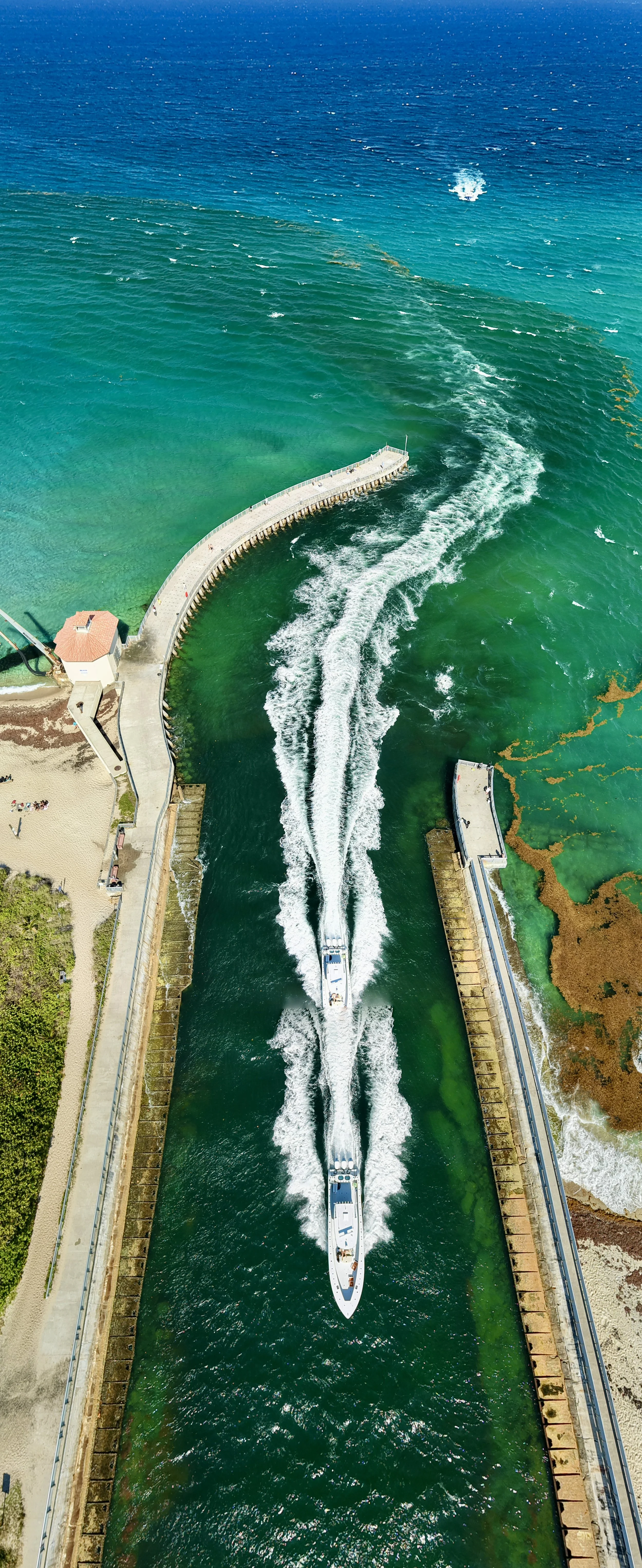 Aerial view of a boat passing through a canal with a curved seawall, clear green water, and a sandy beach with people near the coastline.