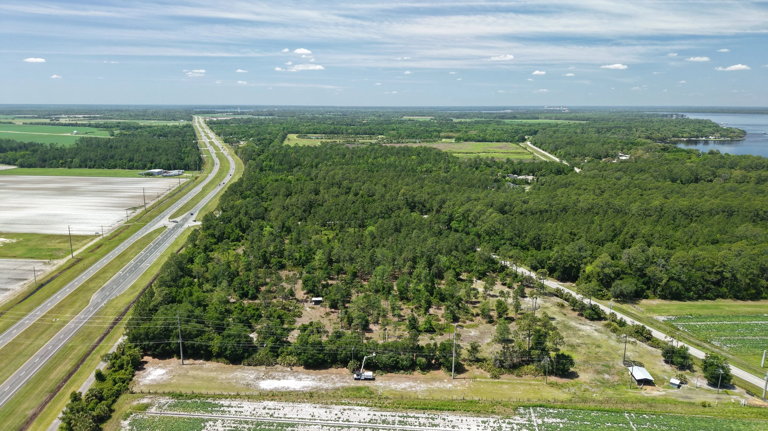 Aerial view of a landscape with a highway, green forest, fields, and a body of water under a partly cloudy sky.