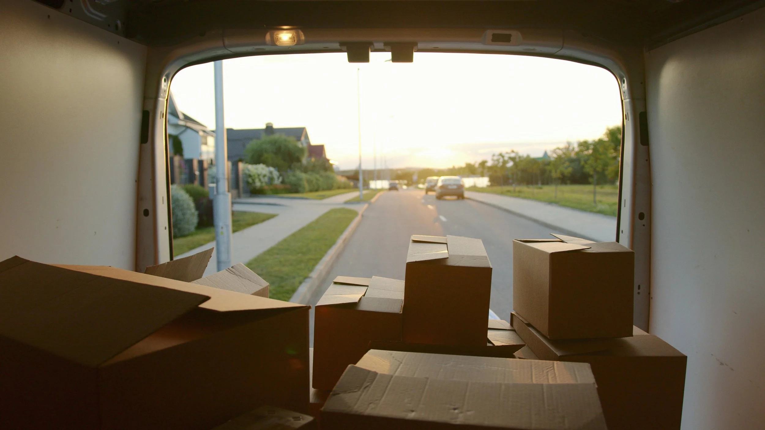 View from inside a vehicle, looking out through an open back door. Several cardboard boxes are inside, with some on the floor and others stacked. Outside, a residential street with houses, trees, and a few cars is visible during sunset.