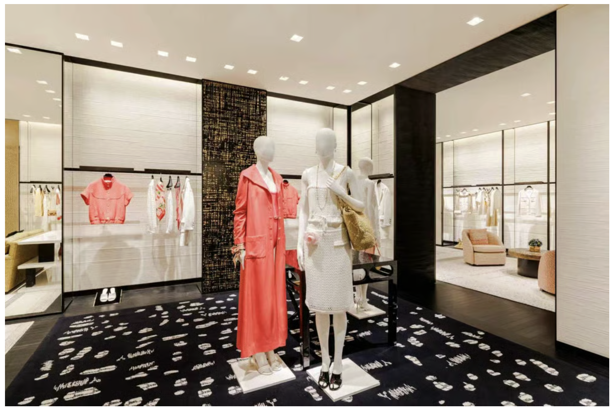 Clothing store with mannequins displaying pink and white clothing, clothing racks with garments, patterned carpet, and a seating area in the background.