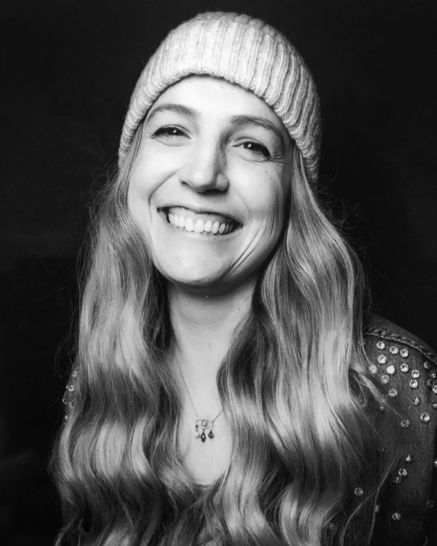 A black-and-white portrait of a smiling woman with long wavy hair wearing a knit beanie and a shirt with a polka dot pattern, also showing a necklace.
