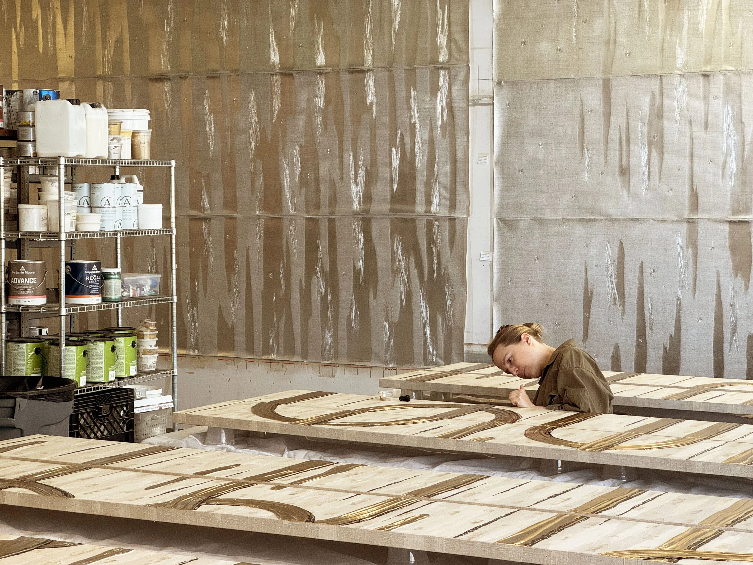 A person working on assembling or painting a large wooden surface, possibly part of furniture or cabinetry, in a workshop with shelves containing paint cans and supplies, and a textured wall in the background.