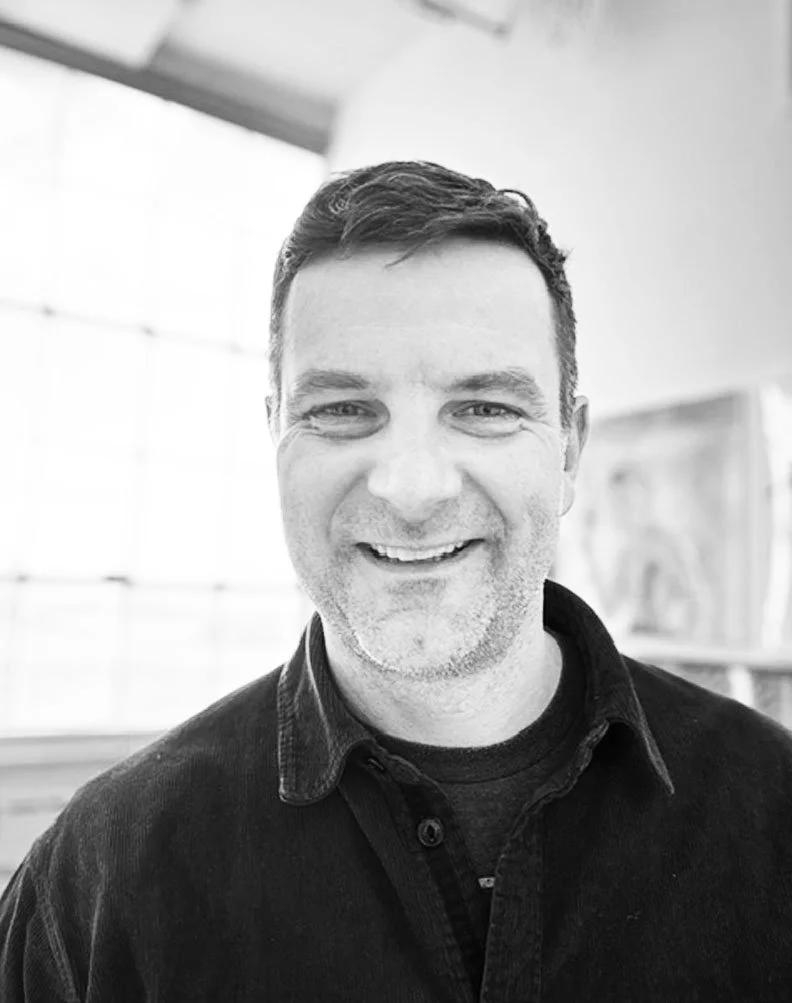 A smiling man with short dark hair and light stubble in an indoor setting with large windows and blurry artwork in the background.