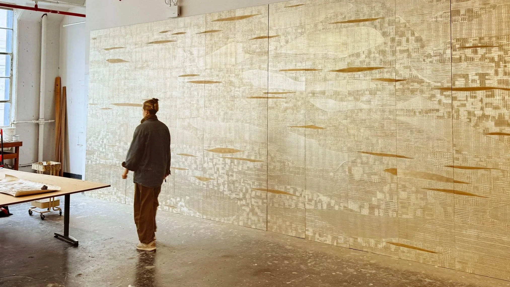 A person with a bun hairstyle and casual clothing standing inside an art studio or workshop, looking at a large, intricate artwork on the wall that resembles a cityscape or landscape with abstract patterns.