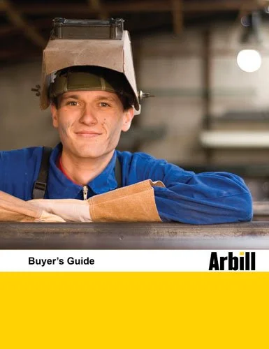 A young man in a blue work uniform wearing a welding helmet pushed back on his head, smiling, leaning on a work surface in a workshop.