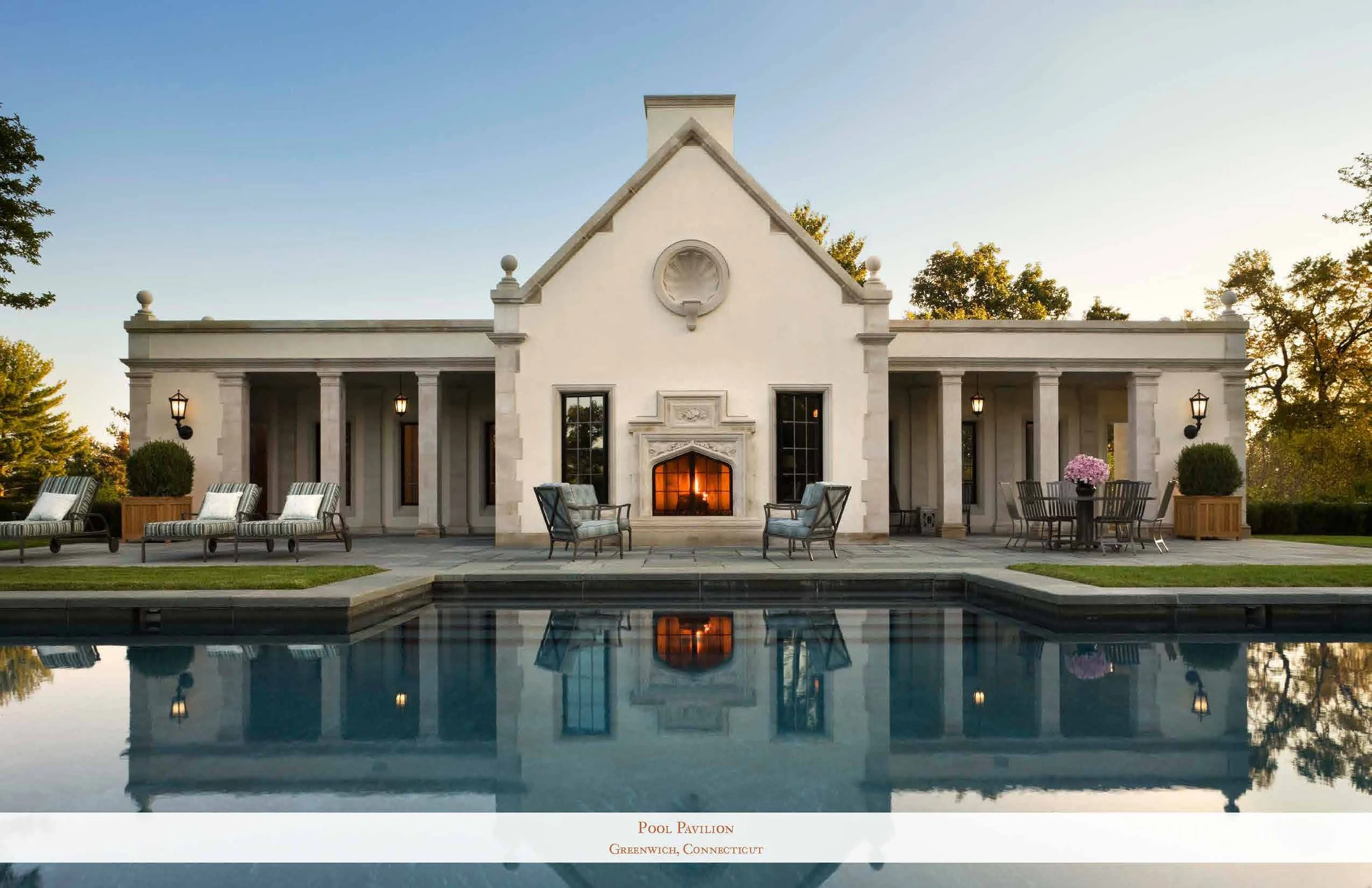 A white pool pavilion with classical columns, a fireplace, and outdoor seating area, reflected in a swimming pool, with trees and a sunset sky in the background.