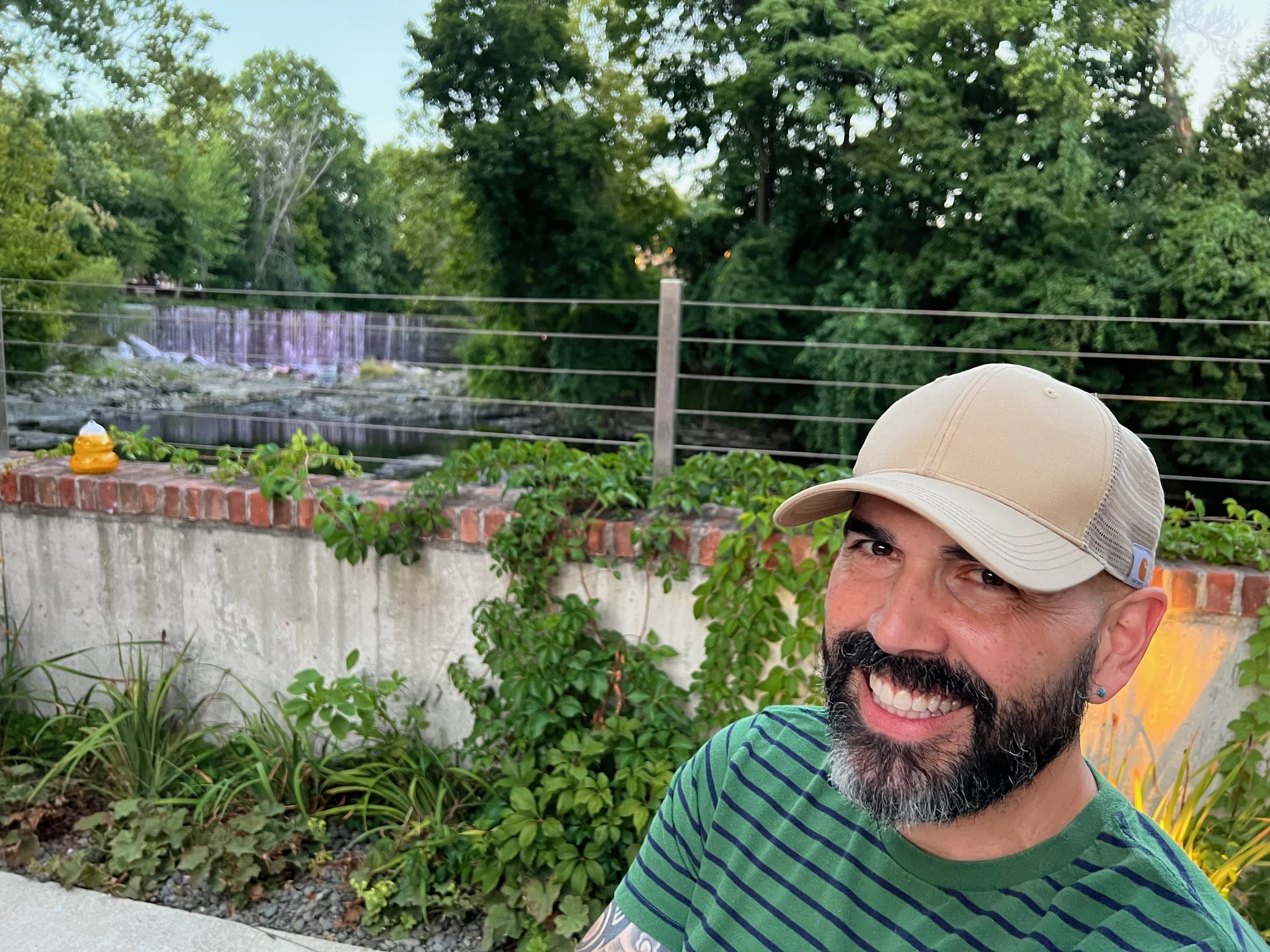 A smiling man with a beard wearing a tan cap and green striped shirt, standing outdoors near plants and a brick wall, with a background of green trees and a water feature.