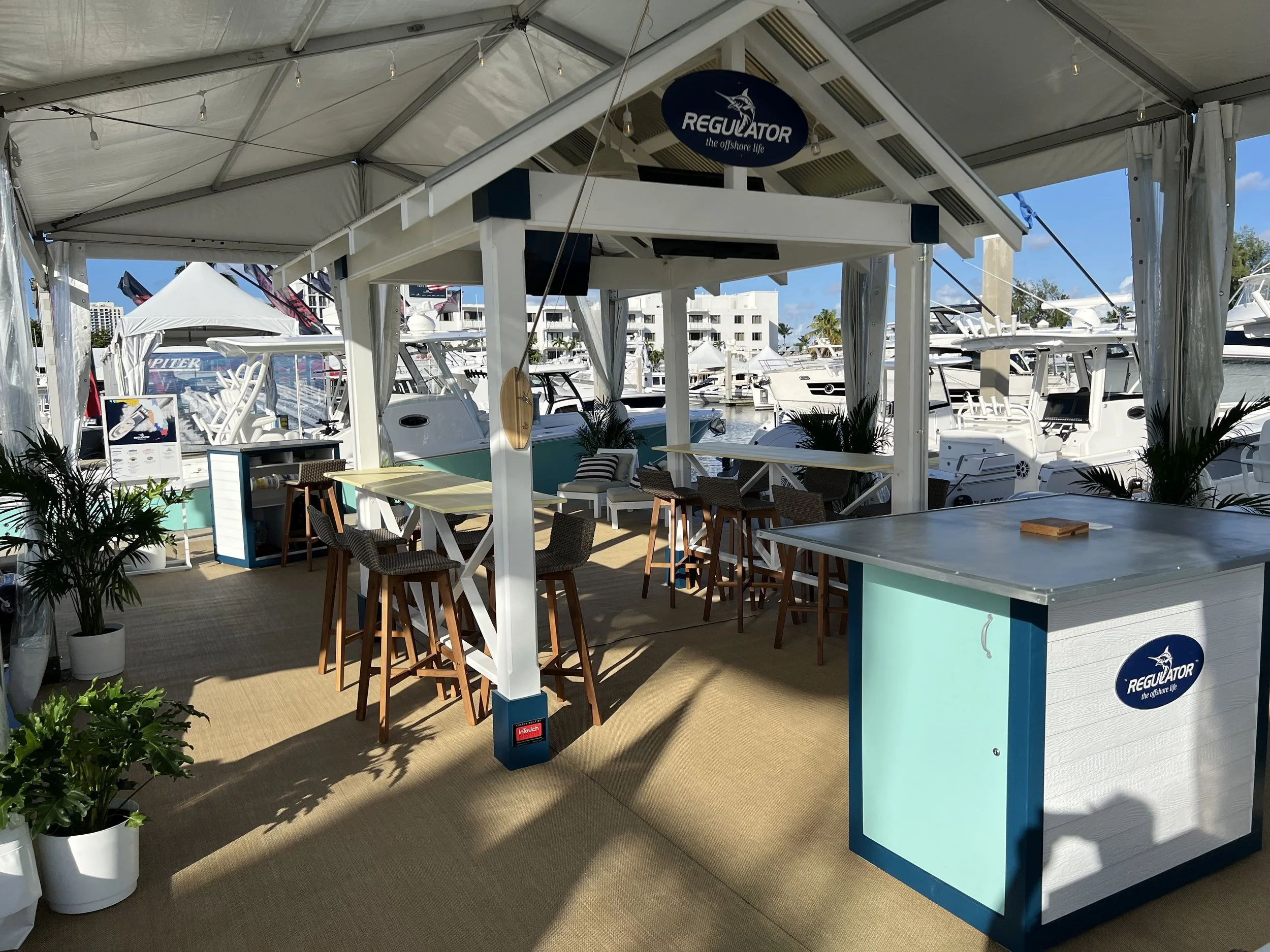 A marina with boats docked, an outdoor dining area under a tent with chairs and tables, and a sign for 'Regulator the offshore life'.