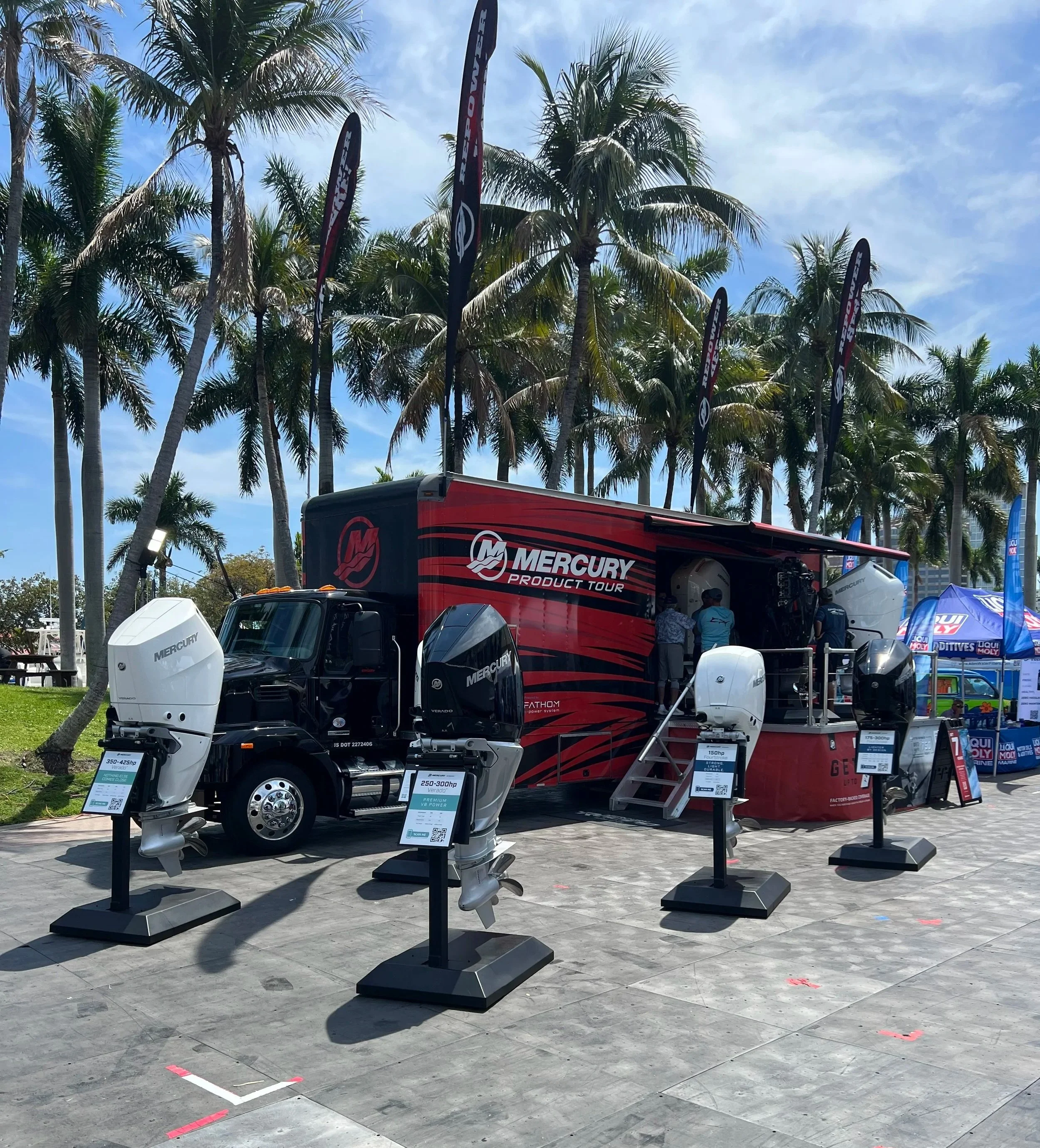 An outdoor display of Mercury marine outboard motors at a boat show, with palm trees in the background under a partly cloudy sky.