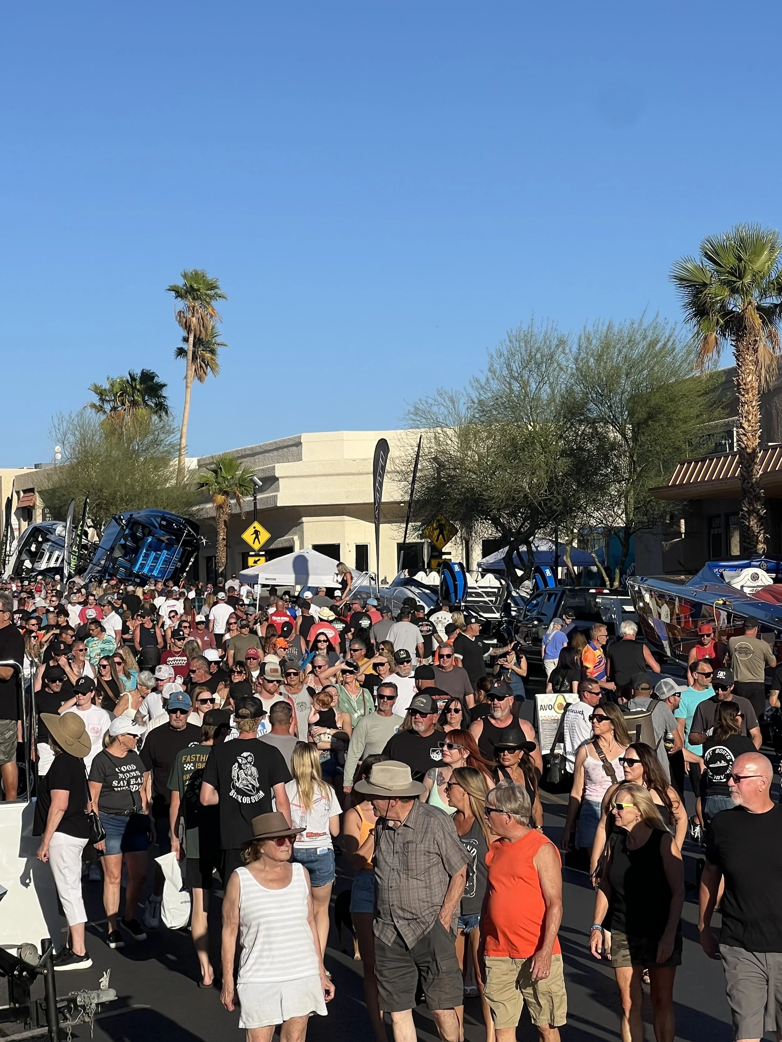 Crowd of people gathering outdoors on a sunny day with palm trees, signage, and tents visible in the background.