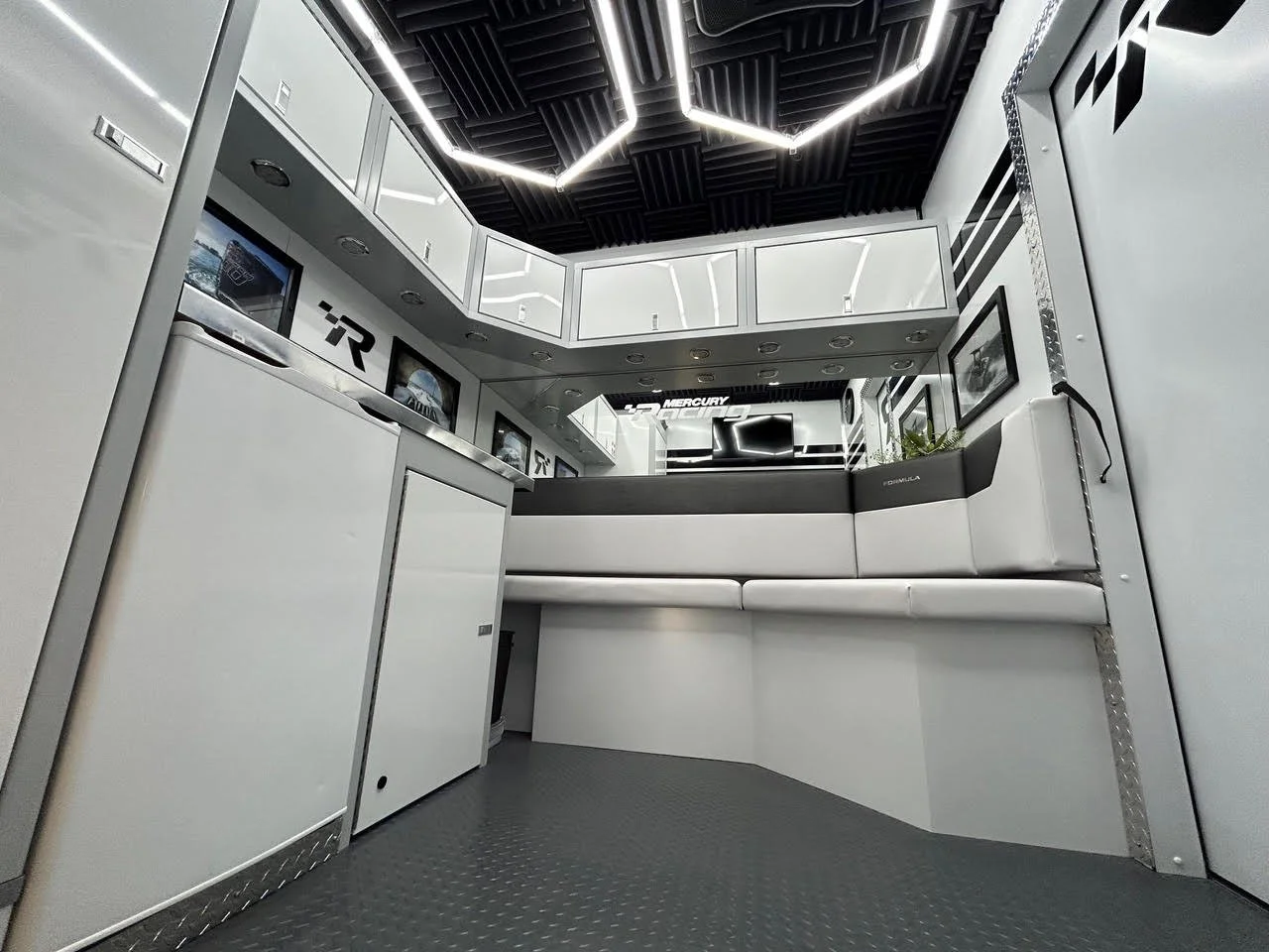 Interior of a modern workspace or lounge area with white cabinetry, framed pictures on the walls, a black ceiling with geometric lighting, and a white cushioned bench along the wall.