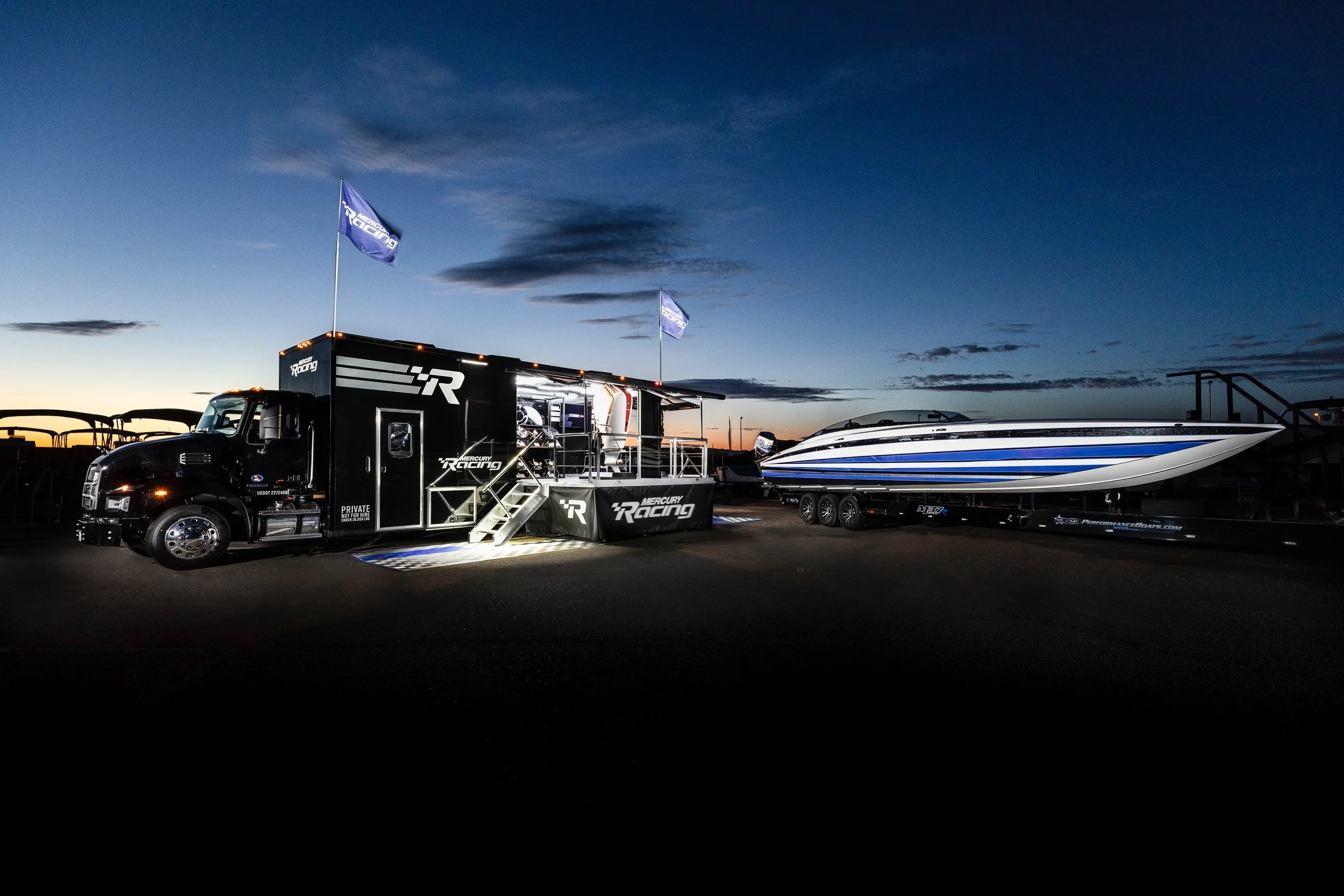 A Mercedes-AMG racing support truck with a boat on a trailer during twilight, with flags and a sky with clouds in the background.