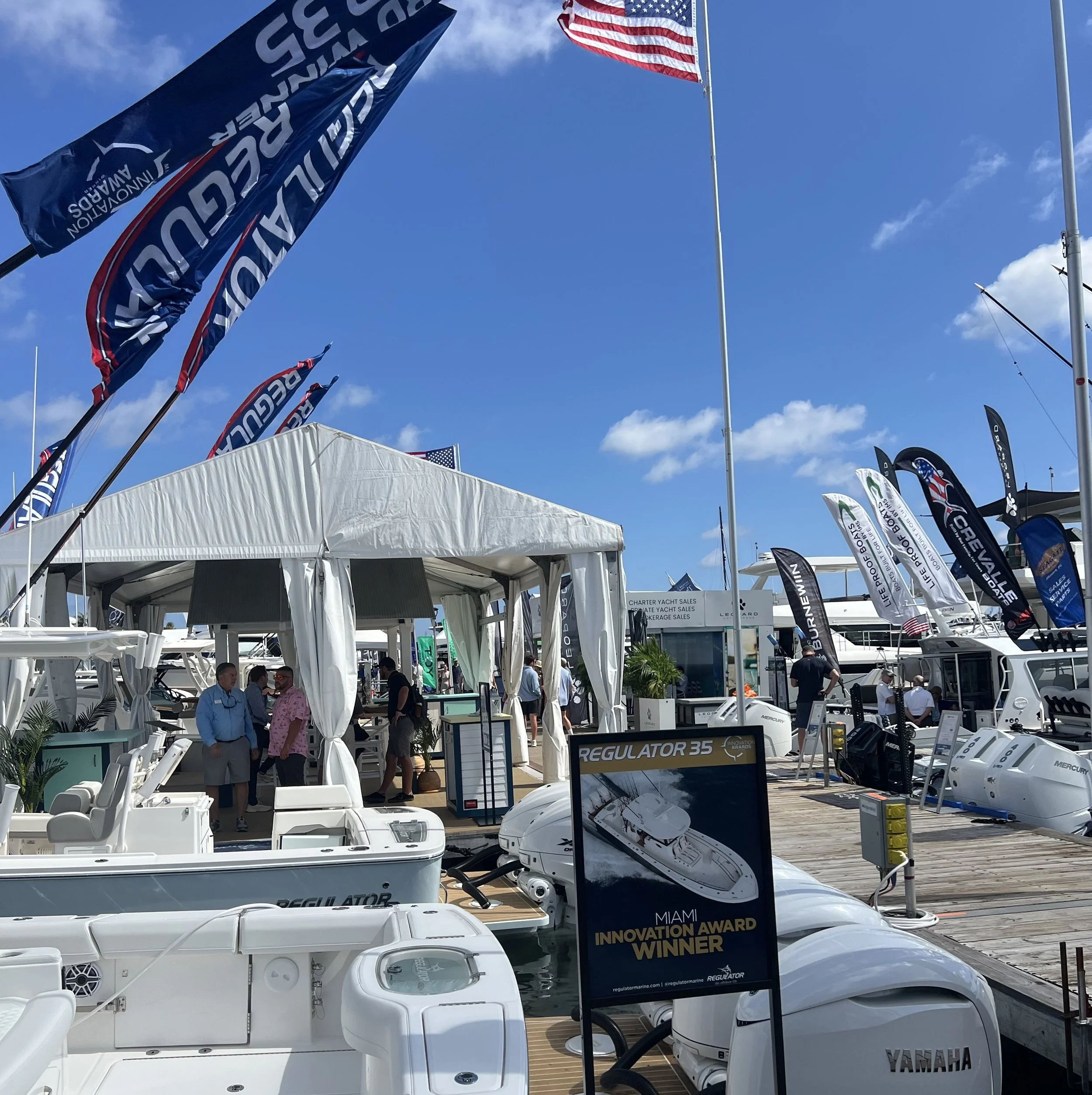 Display of various boats and boat engines at a marine exhibition with flags and people walking around on a bright, partly cloudy day.