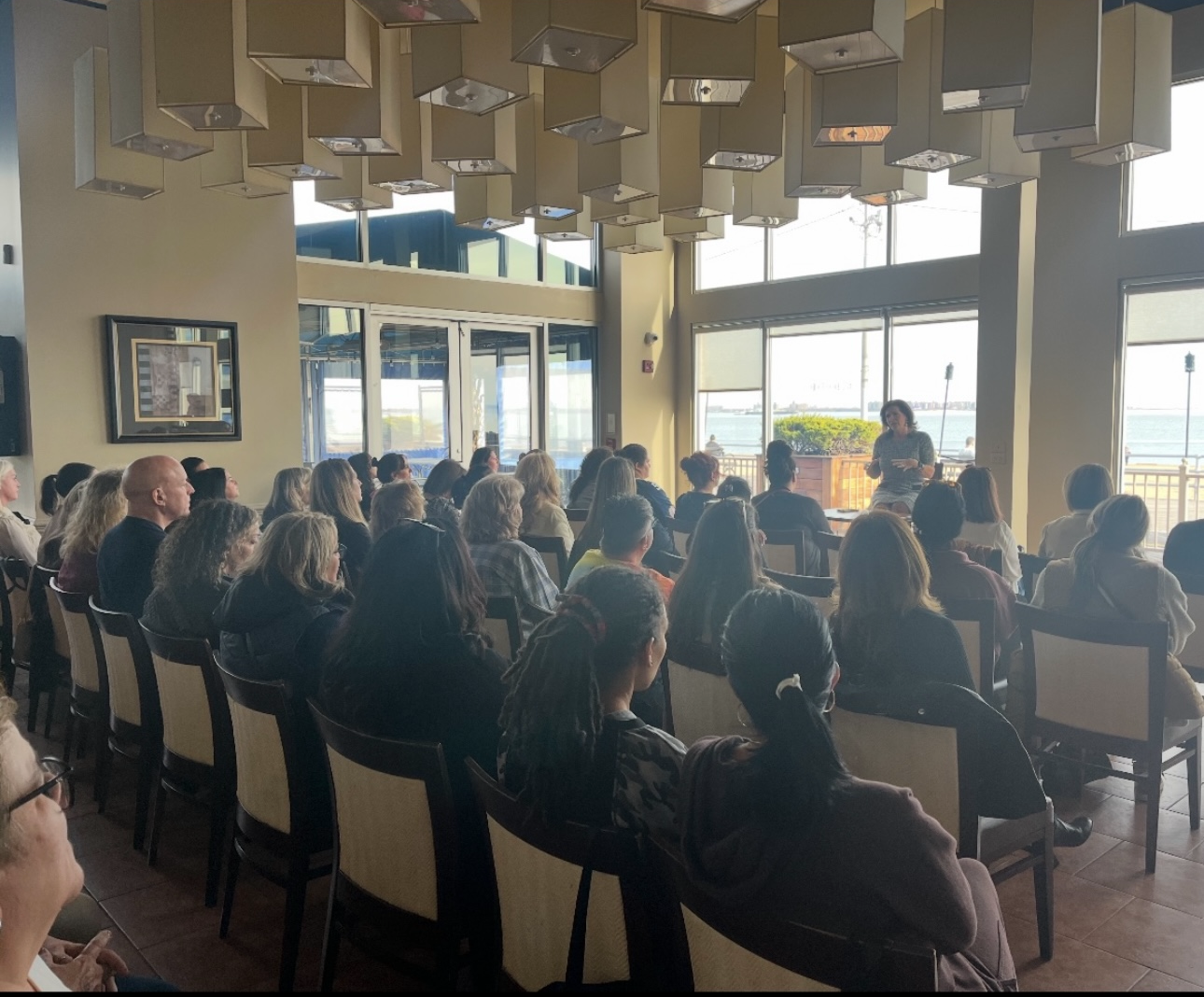A woman giving a presentation to an audience in a room with large windows overlooking a body of water. The audience is seated and attentively listening.