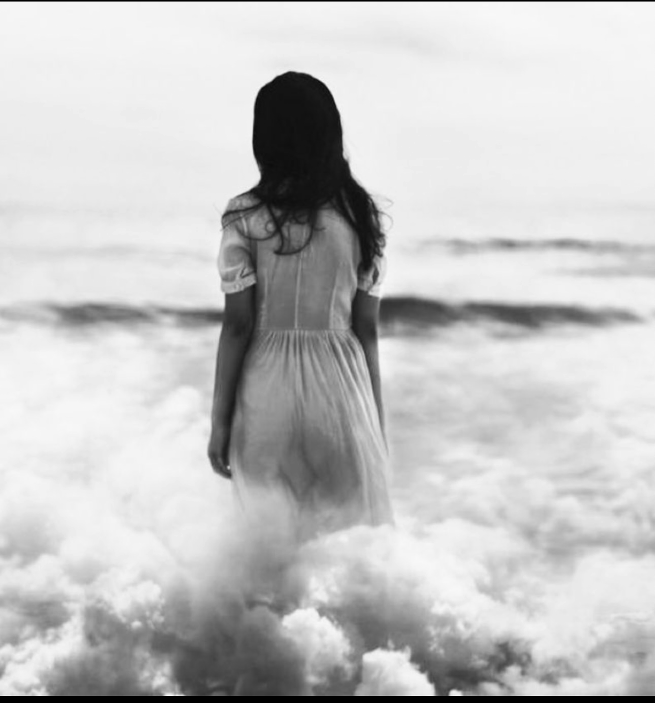 A girl in a dress is standing in the water at the beach, facing away from the camera, looking towards the ocean.