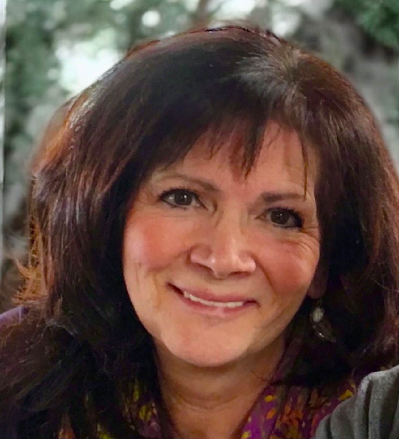 Close-up of a woman with shoulder-length dark brown hair, smiling, wearing earrings and a colorful patterned blouse, with a blurred outdoor background.