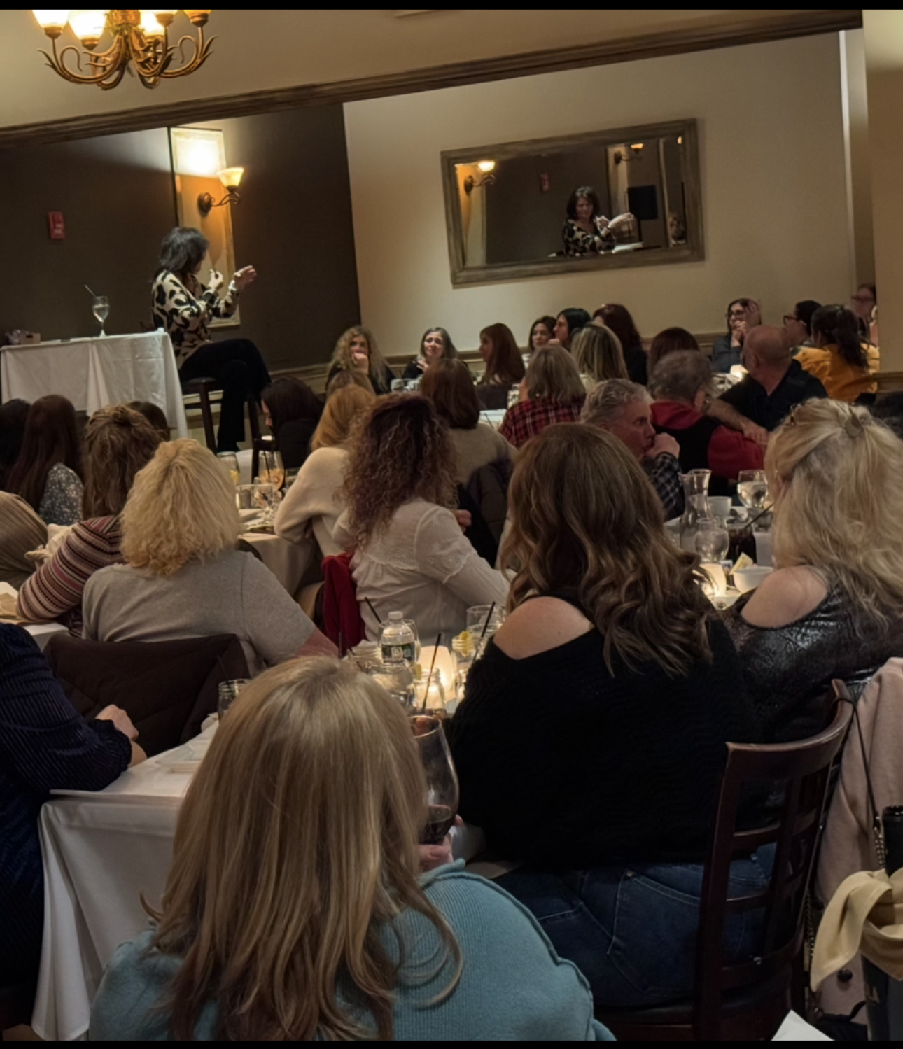 A woman with short dark hair wearing a leopard print top is speaking at a microphone on a small stage to an audience seated at tables inside a banquet hall. The audience consists mainly of women, some with curly and straight hair, wearing casual and semi-formal clothing. Some tables have water bottles, glasses, and candles, and there's a large mirror on the wall behind the speaker.