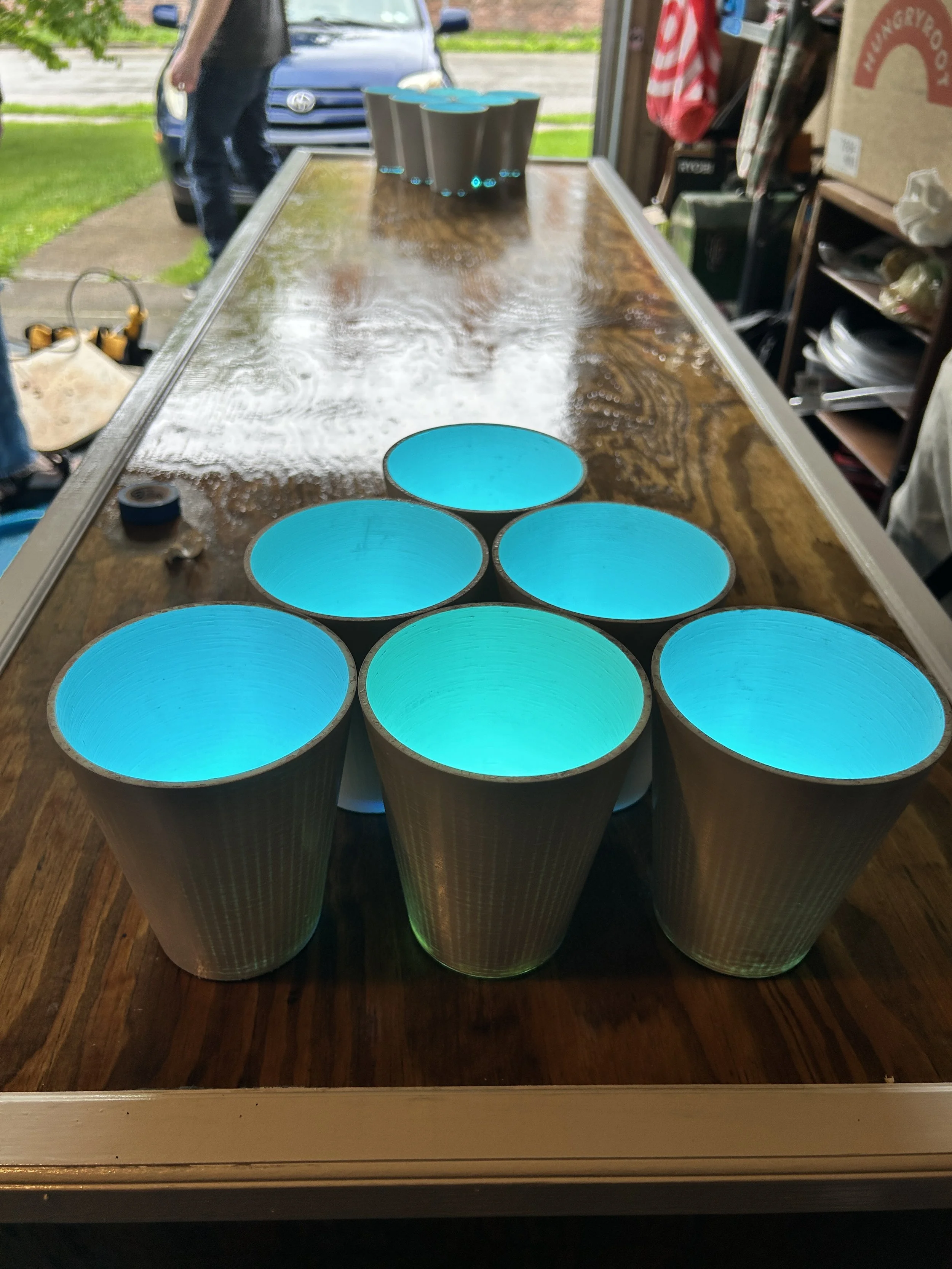A row of six illuminated blue LED illuminated tubes viewed from the front, placed on a wooden table inside a store or workshop, with a garage door and outdoor scenery visible in the background.