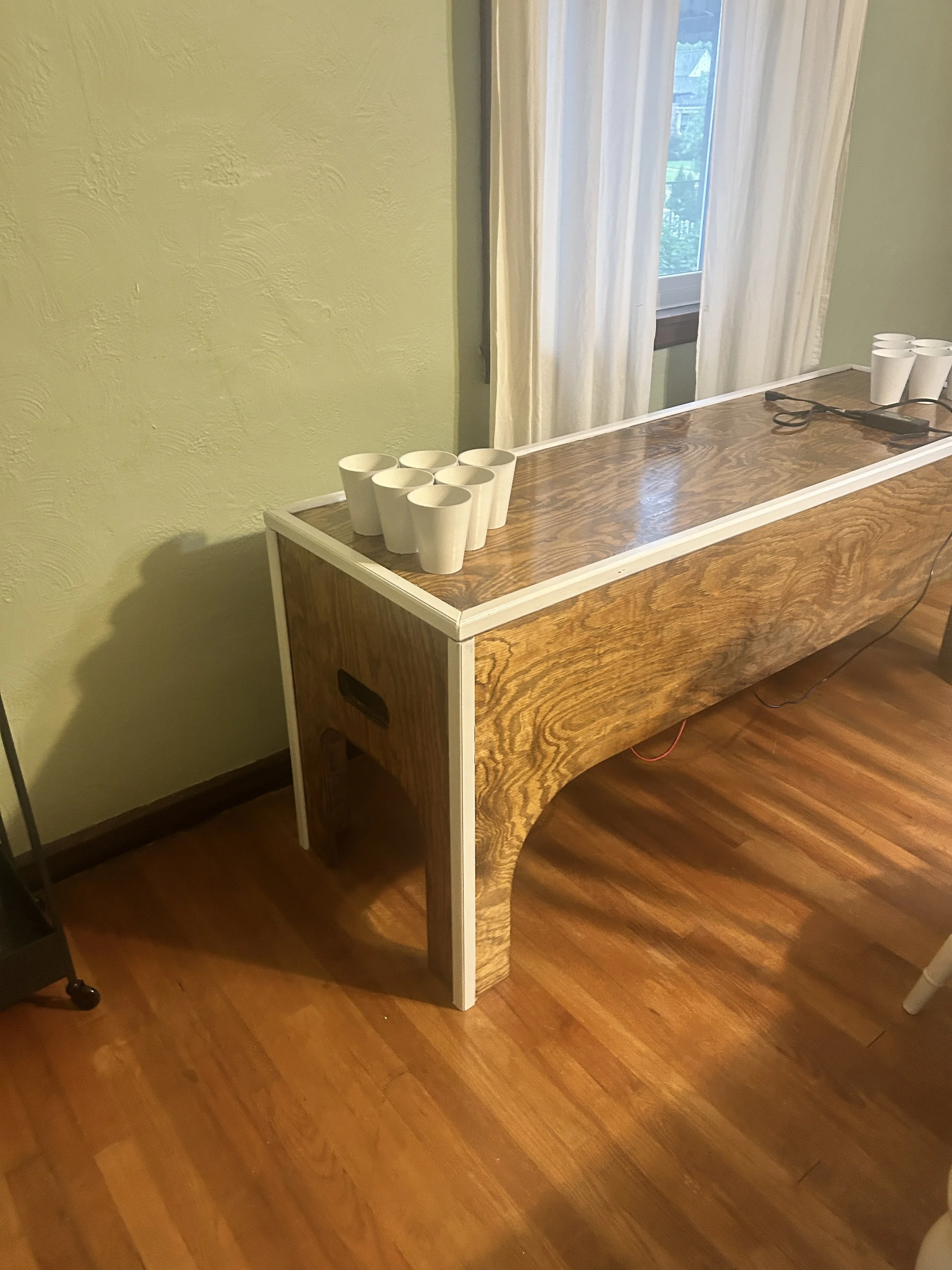 A wooden table with white cups on top, located near a window with white curtains, in a room with green walls and wooden flooring.