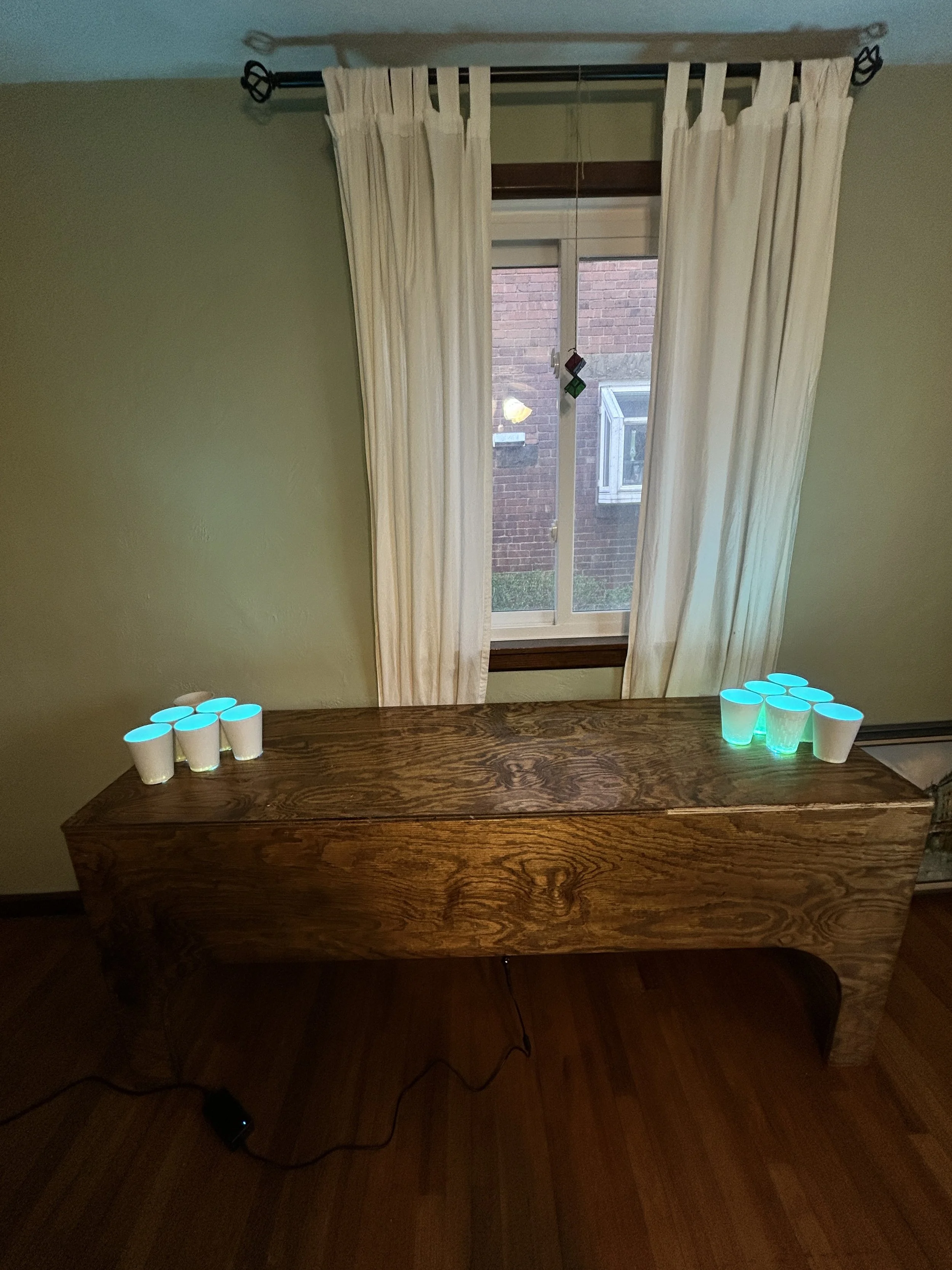 A wooden table with eight glowing cups, four on each side, placed near a window with cream curtains, a brick wall, and a small window outside. An electrical cord is visible on the floor.