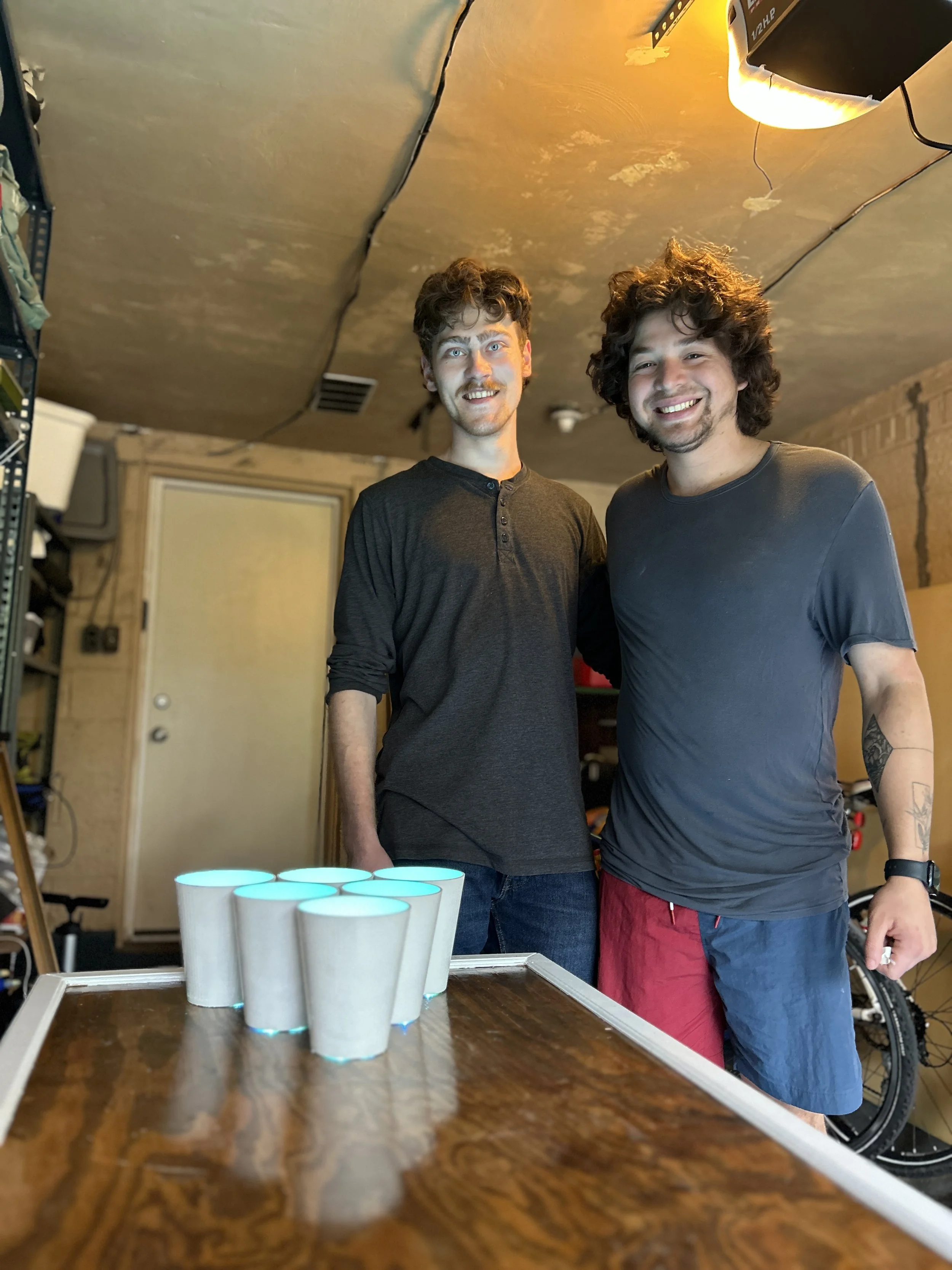 Two young men smiling and standing behind a table with six cups that have glowing blue lights at the bottom. One man has curly hair and a beard, wearing a dark t-shirt, and the other has shaggy hair and a beard, wearing a dark shirt and red and blue shorts.